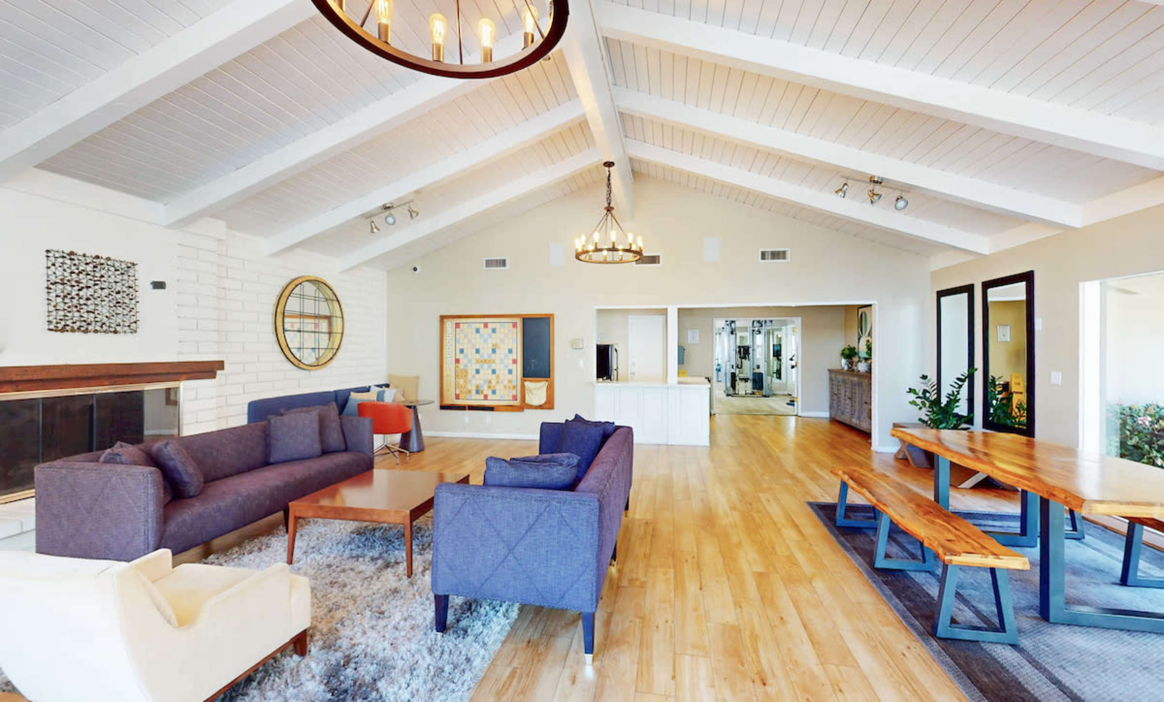 The image shows a spacious living room with a high ceiling, featuring a mix of seating options, a large dining table, and an open layout connecting to a kitchen area.