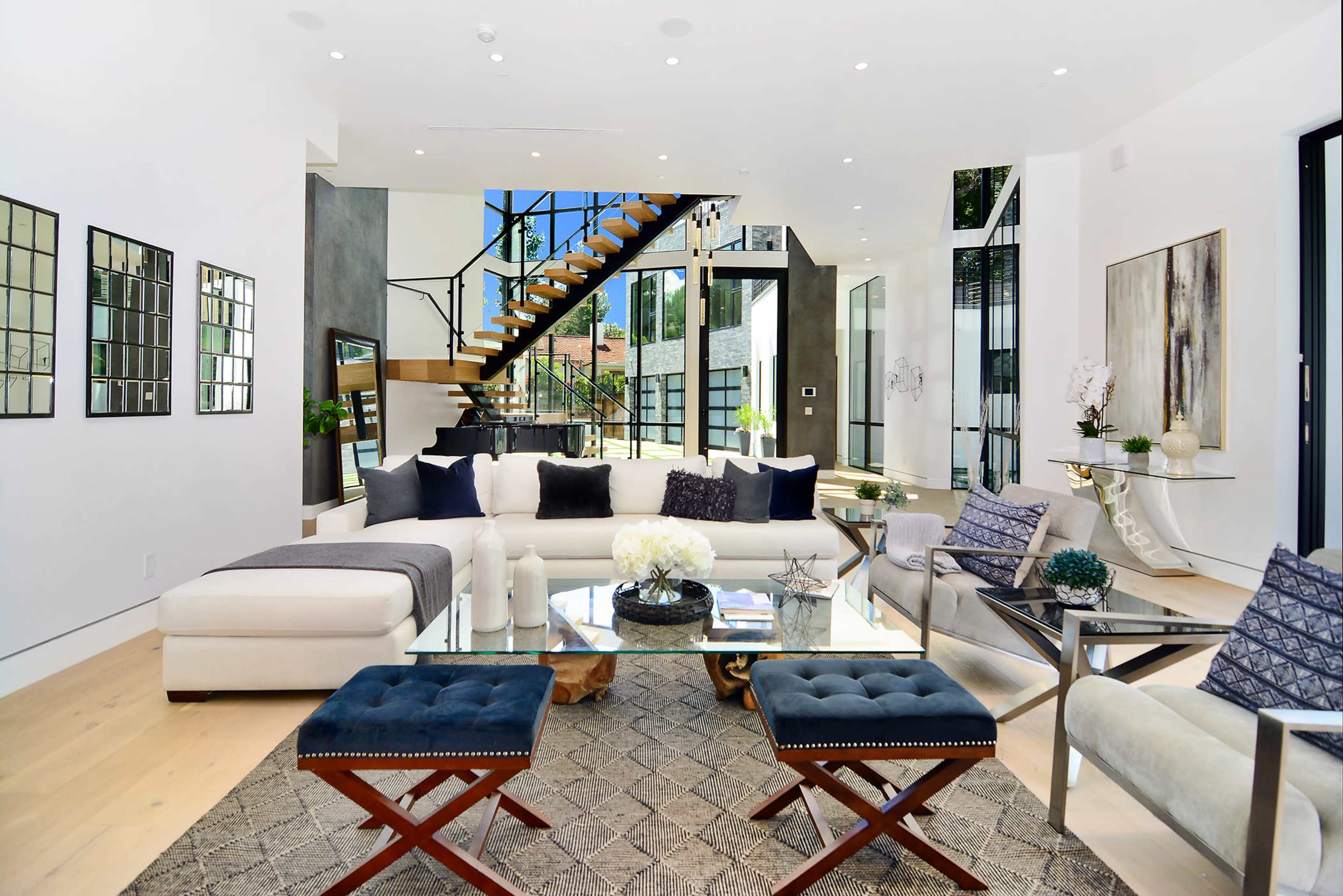 The living room features a modern sectional sofa, a glass coffee table, and a staircase leading to an upper level, with large windows allowing natural light to enter.