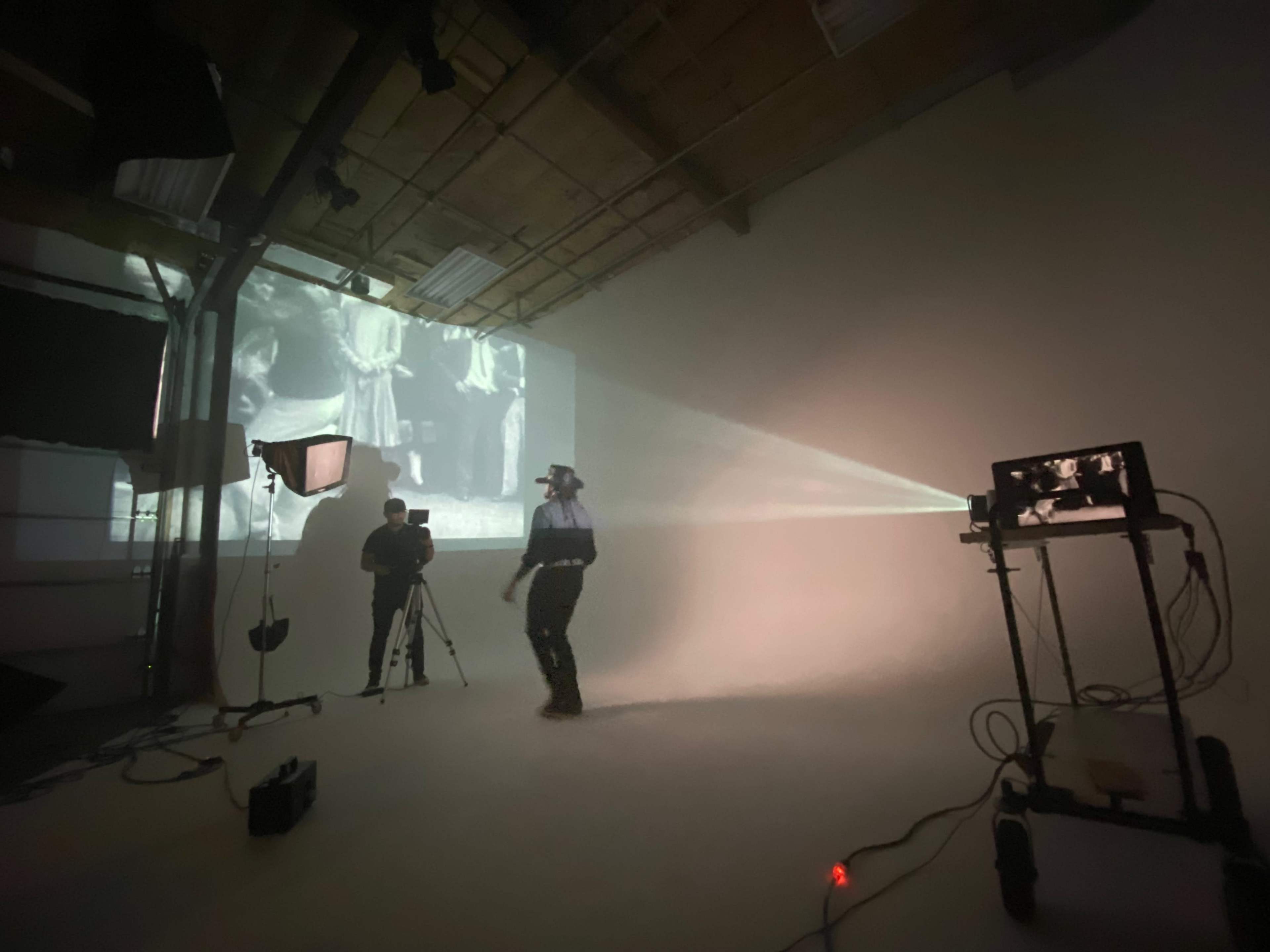 A person operates a camera while another walks in front of a projected black-and-white film in a studio space.