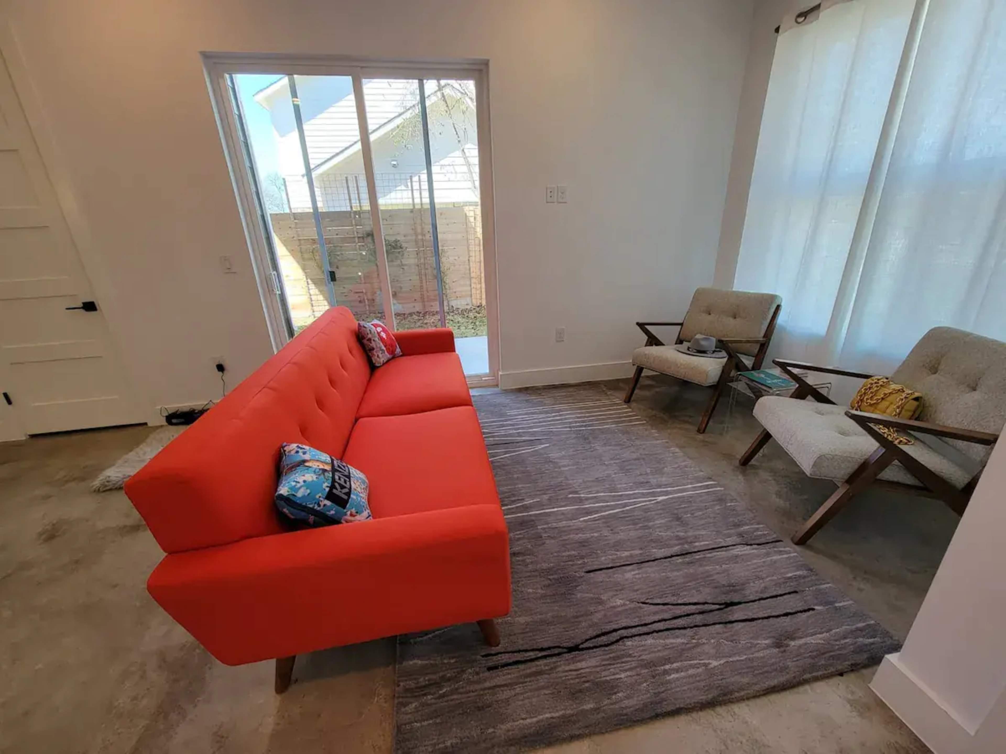 A bright living room features a red sofa facing two light-colored armchairs, with a rug on a concrete floor and natural light coming through a window.