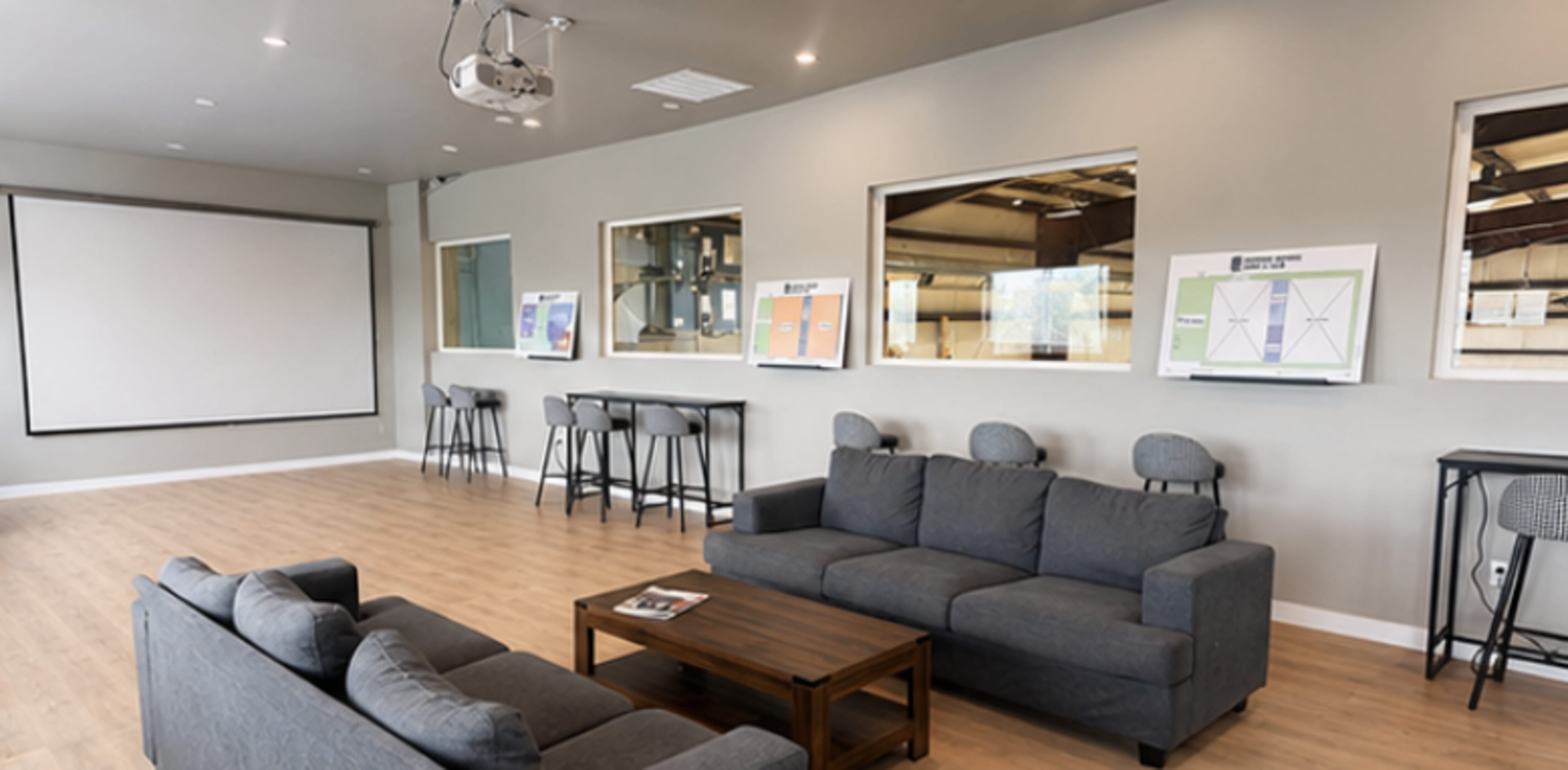 The image shows a modern meeting room featuring gray sofas, a wooden coffee table, and a large projection screen on one wall.