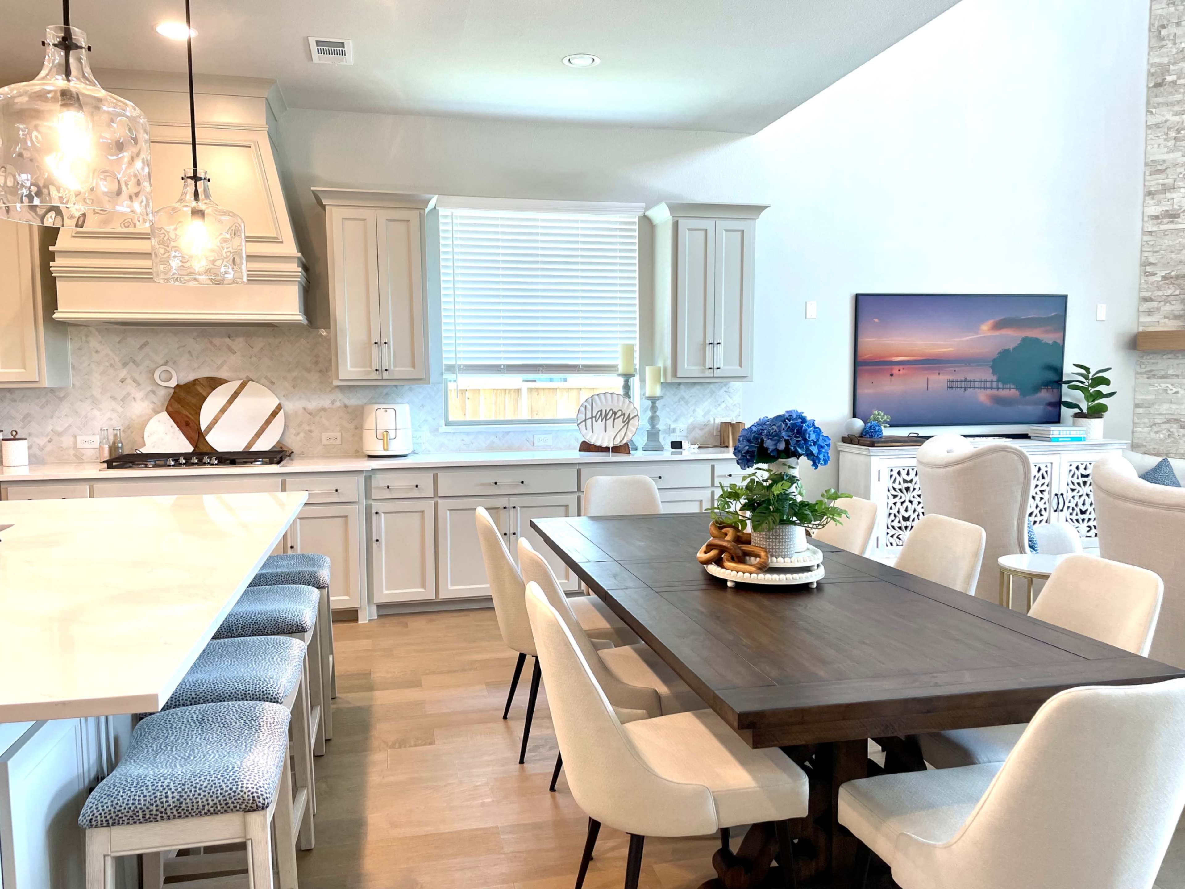 A modern kitchen and dining area with a large wooden table, light-colored cabinetry, and a television mounted on the wall.