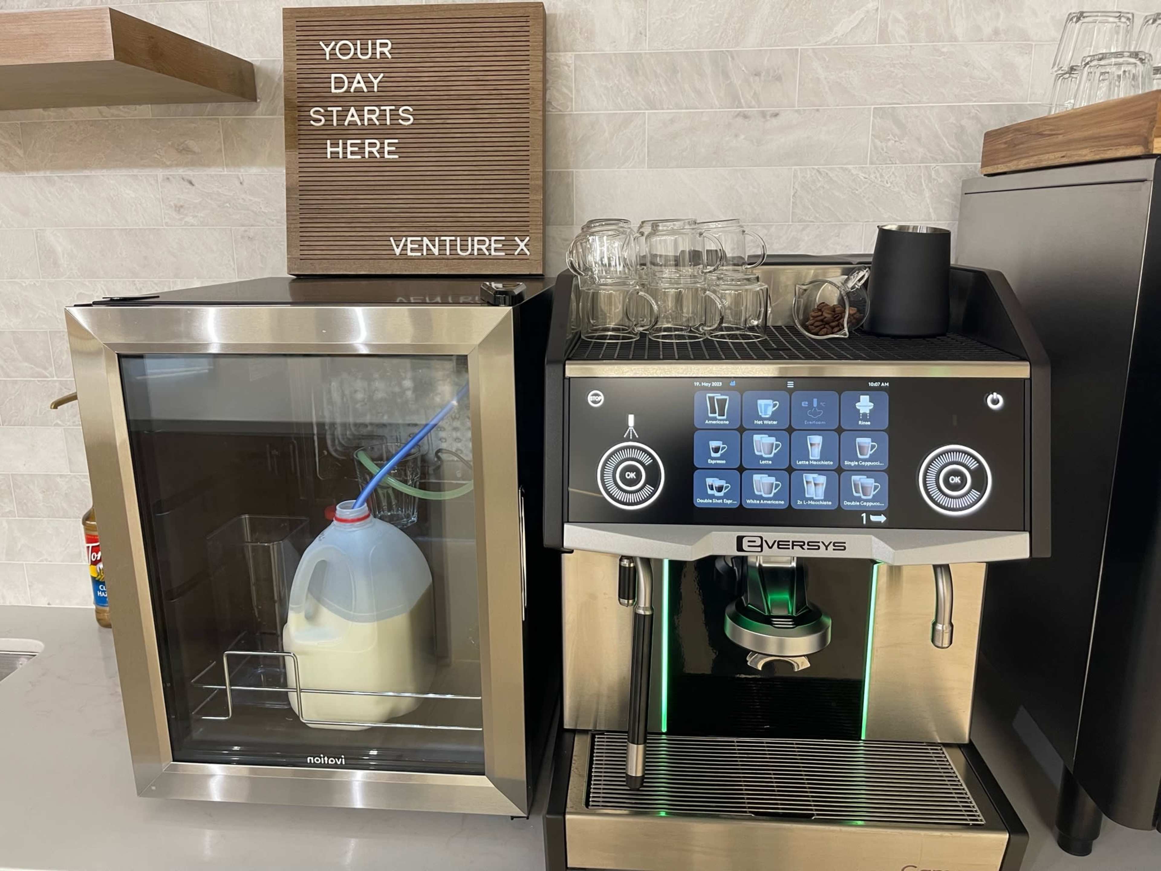 A milk dispenser and a coffee machine are placed side by side on a counter with a sign above that reads "YOUR DAY STARTS HERE" and "VENTURE X."