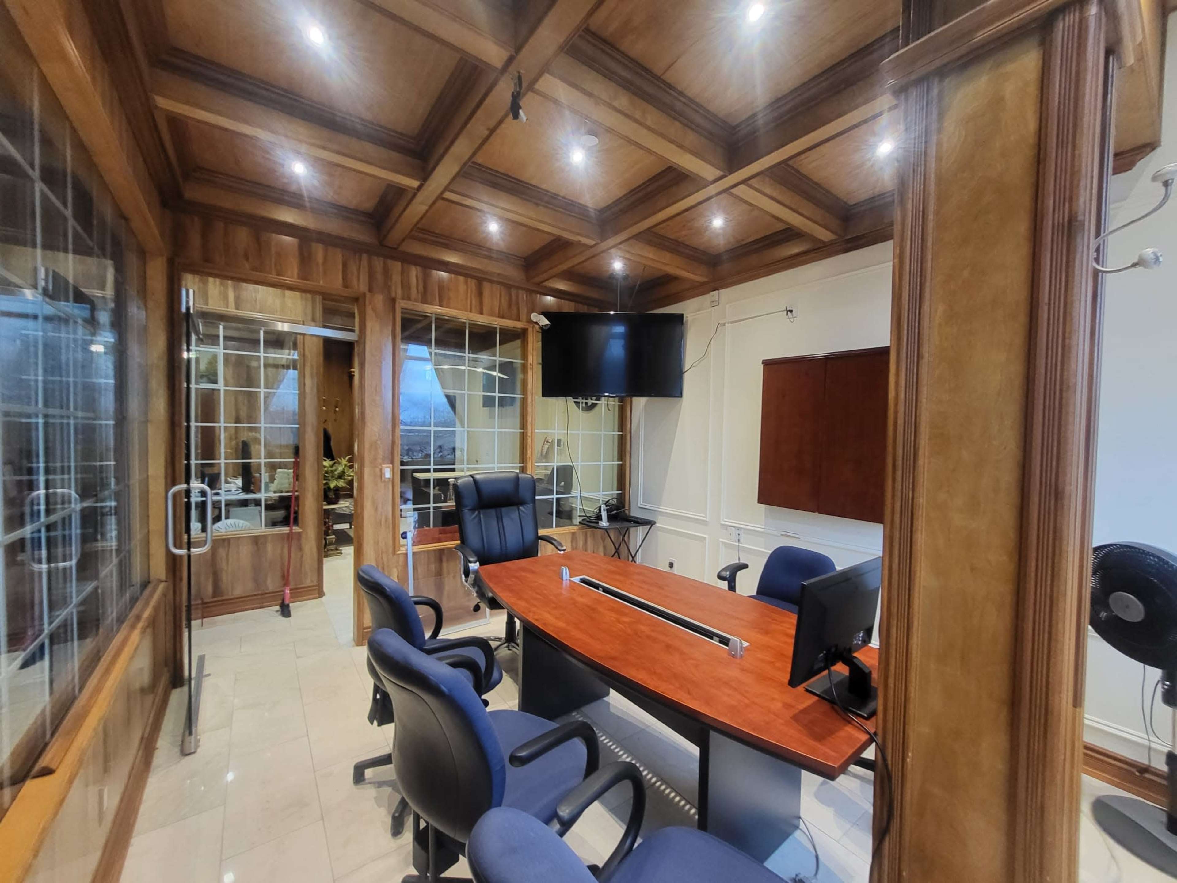 The image shows a well-furnished office with a large wooden conference table, rolling chairs, and a wall-mounted television.