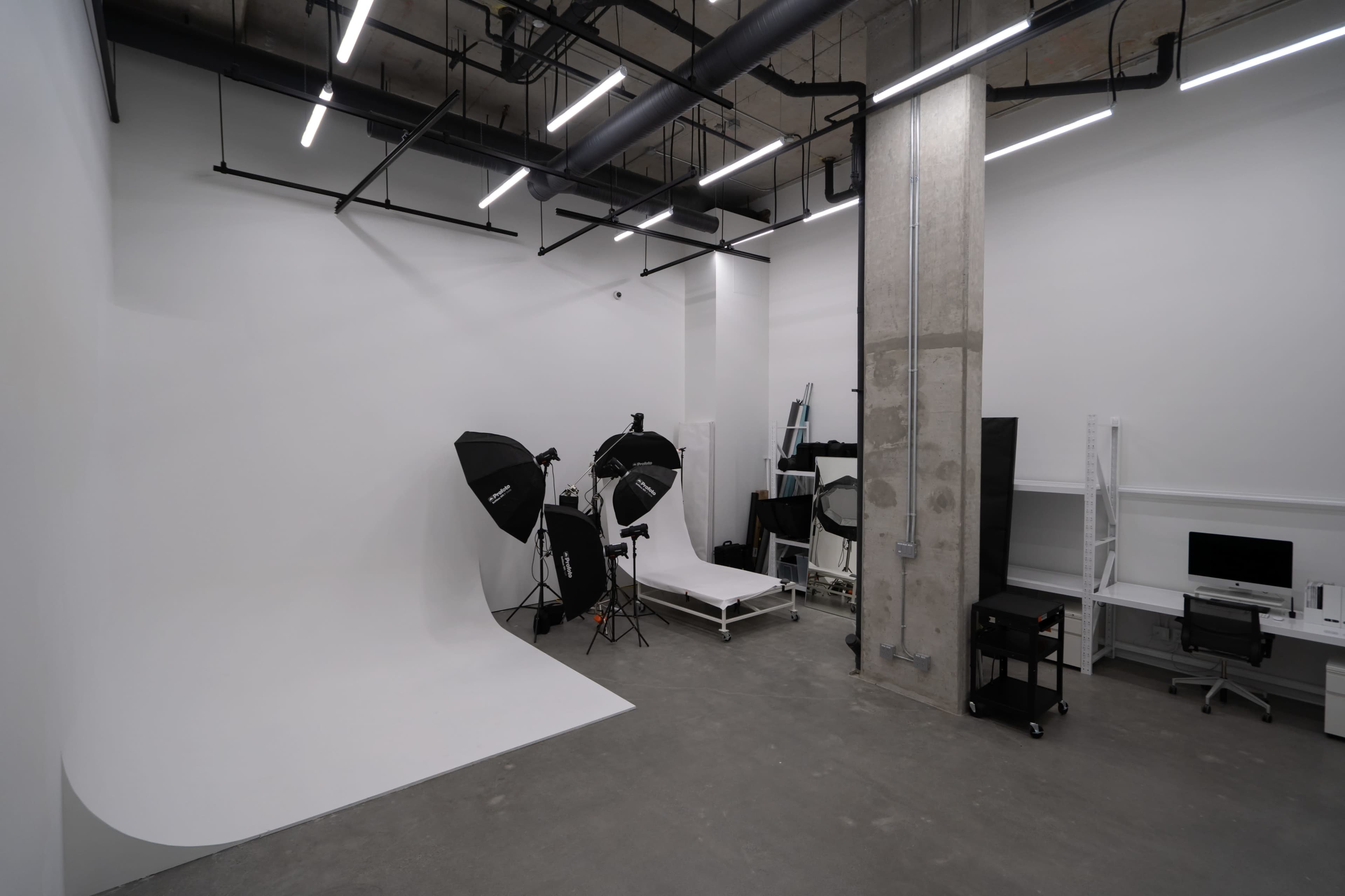 A minimalist photography studio featuring a white backdrop, lighting equipment, a rolling cart, and a workstation.