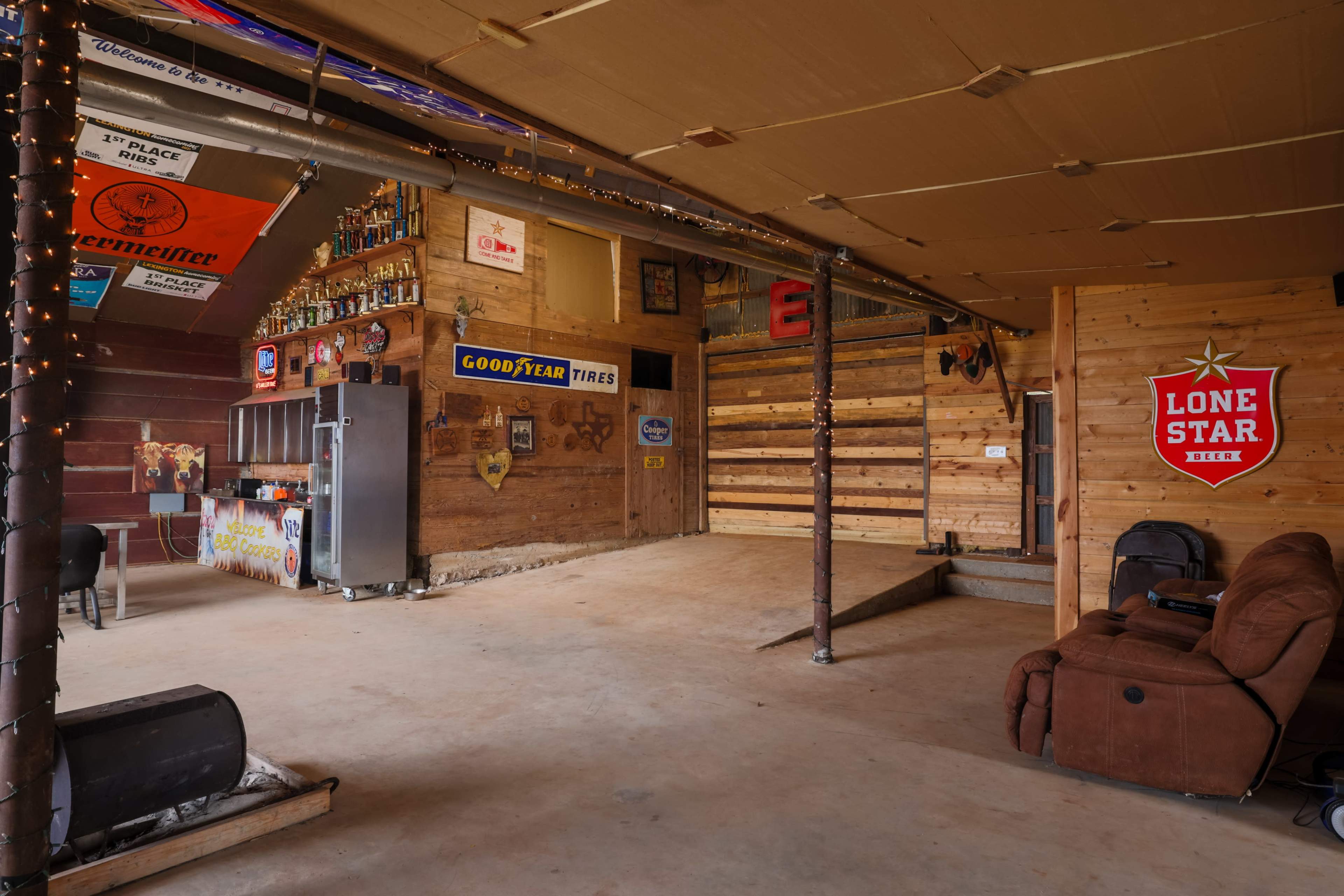 The interior of a rustic garage features wooden walls adorned with vintage signs, a bar area stocked with bottles, and a brown recliner.