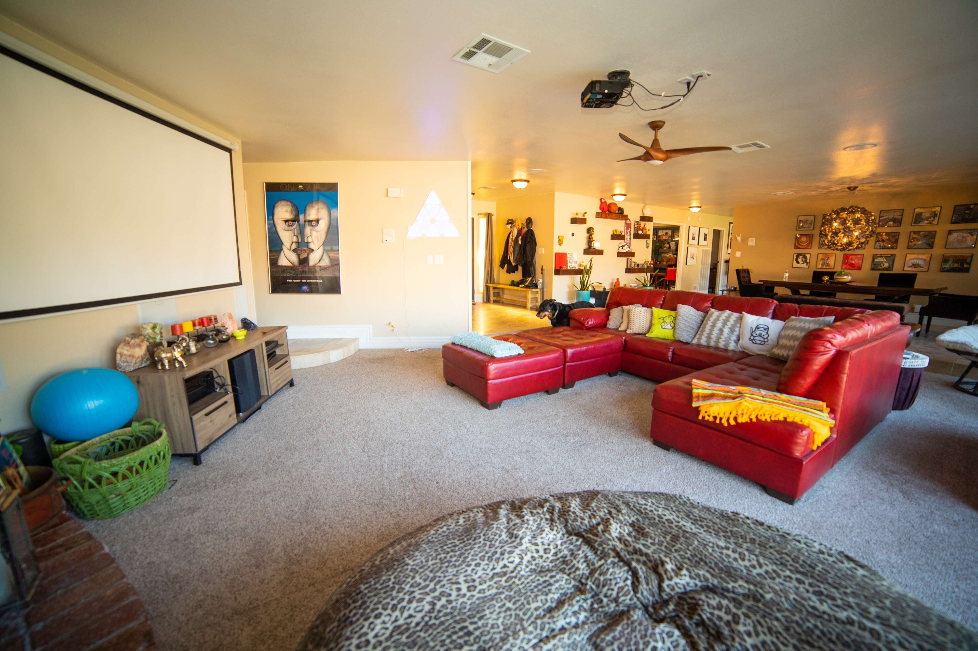 A spacious living room with a large red sectional sofa, a projector screen on the wall, and various decorations, including a poster and a round ottoman.