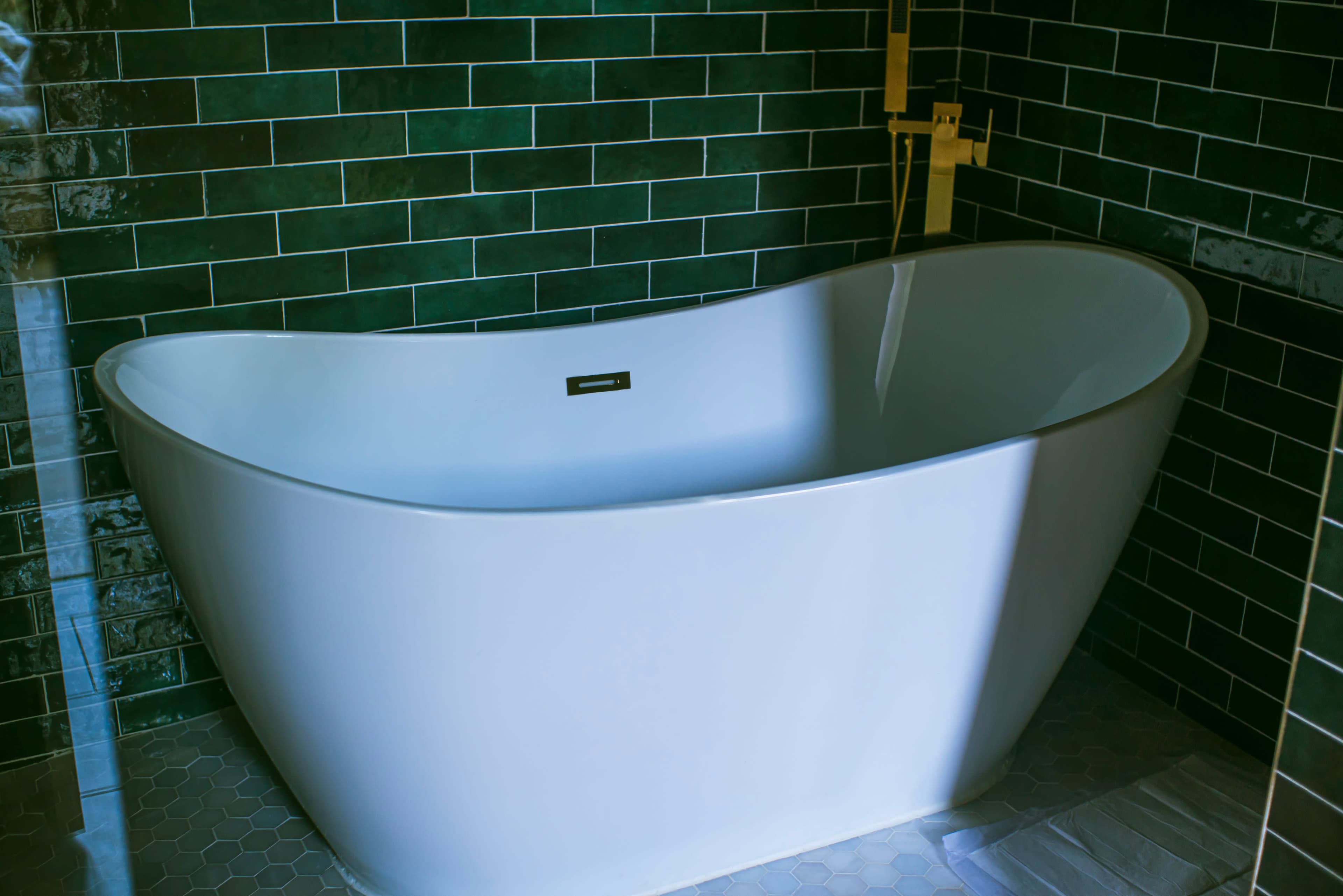 The image features a modern freestanding white bathtub set against a backdrop of dark green tiled walls.