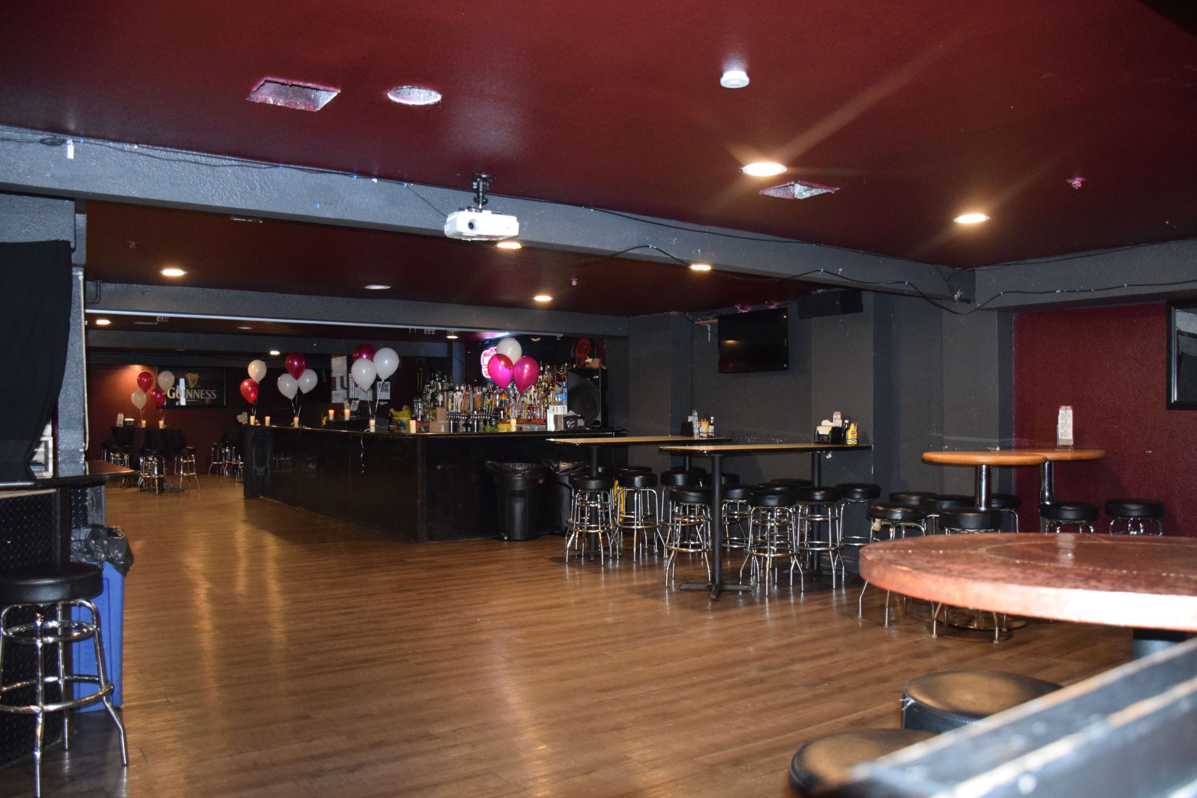 An empty bar area with dark wooden floors, high-top tables, and a long counter lined with chairs, decorated with balloons.