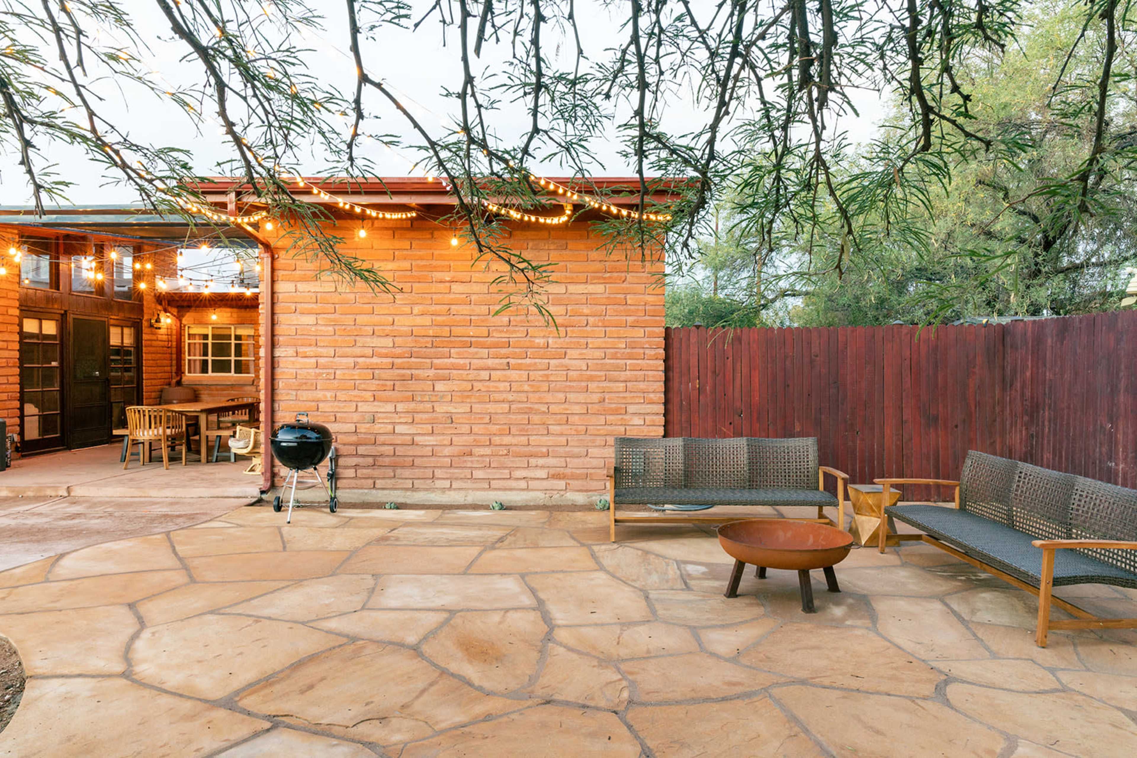 A backyard patio with stone flooring, a charcoal grill, wooden seating, and string lights suspended over a brick wall.