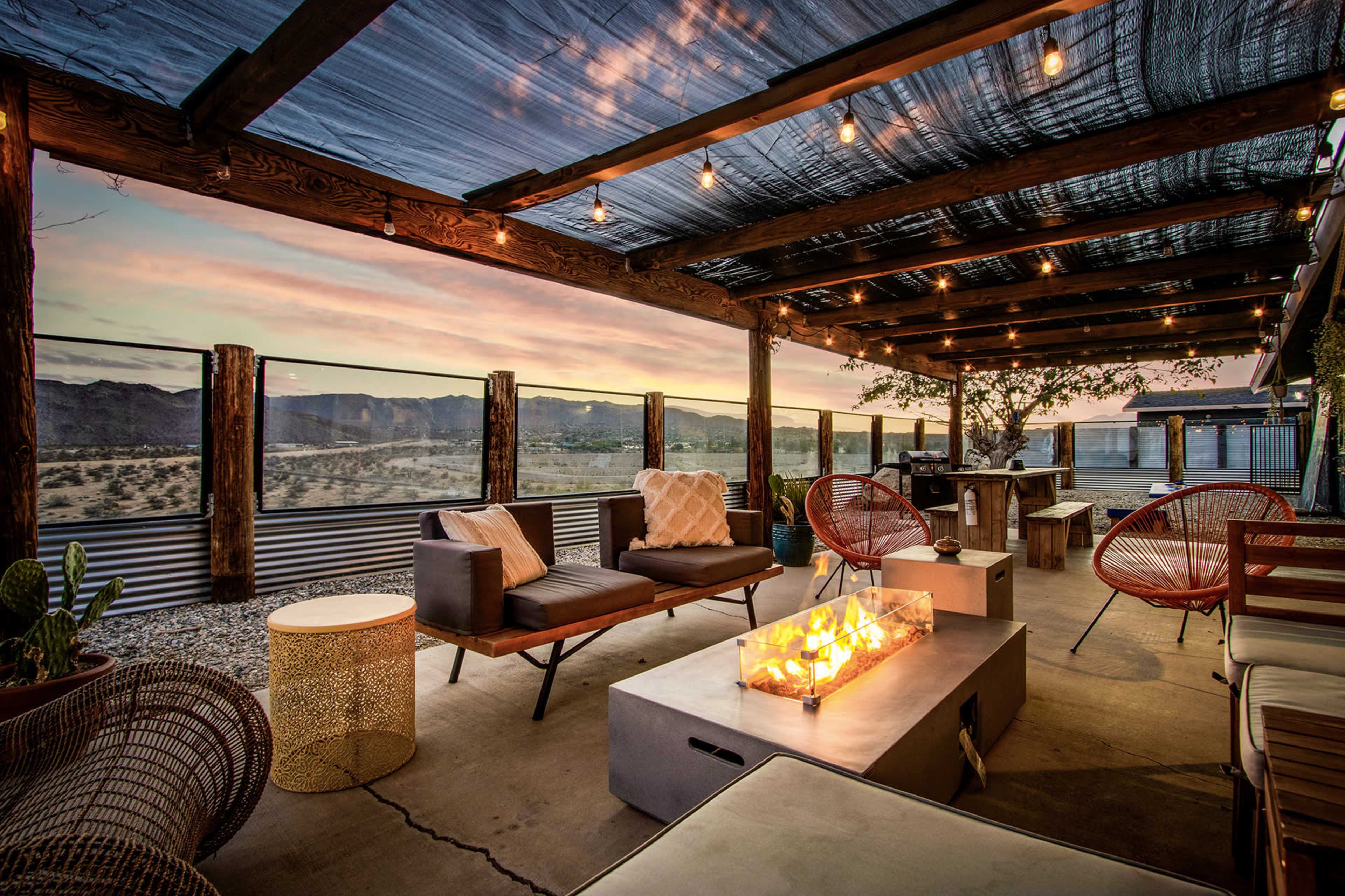 A cozy outdoor patio features modern seating and a fire pit, with a backdrop of a desert landscape and a colorful sunset.