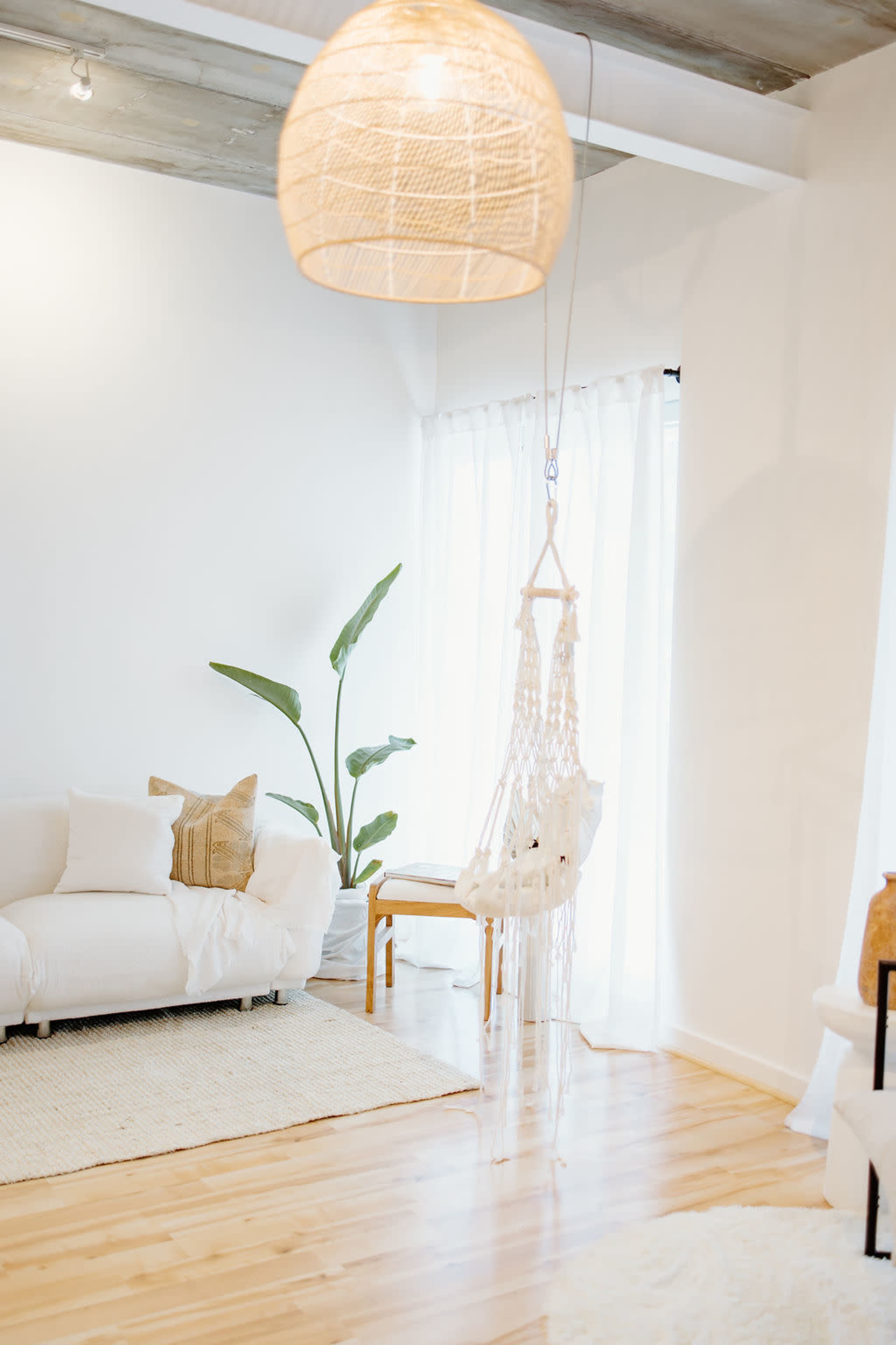 A bright, airy living space featuring a white sofa, a macramé hanging chair, and a large plant near a large window.