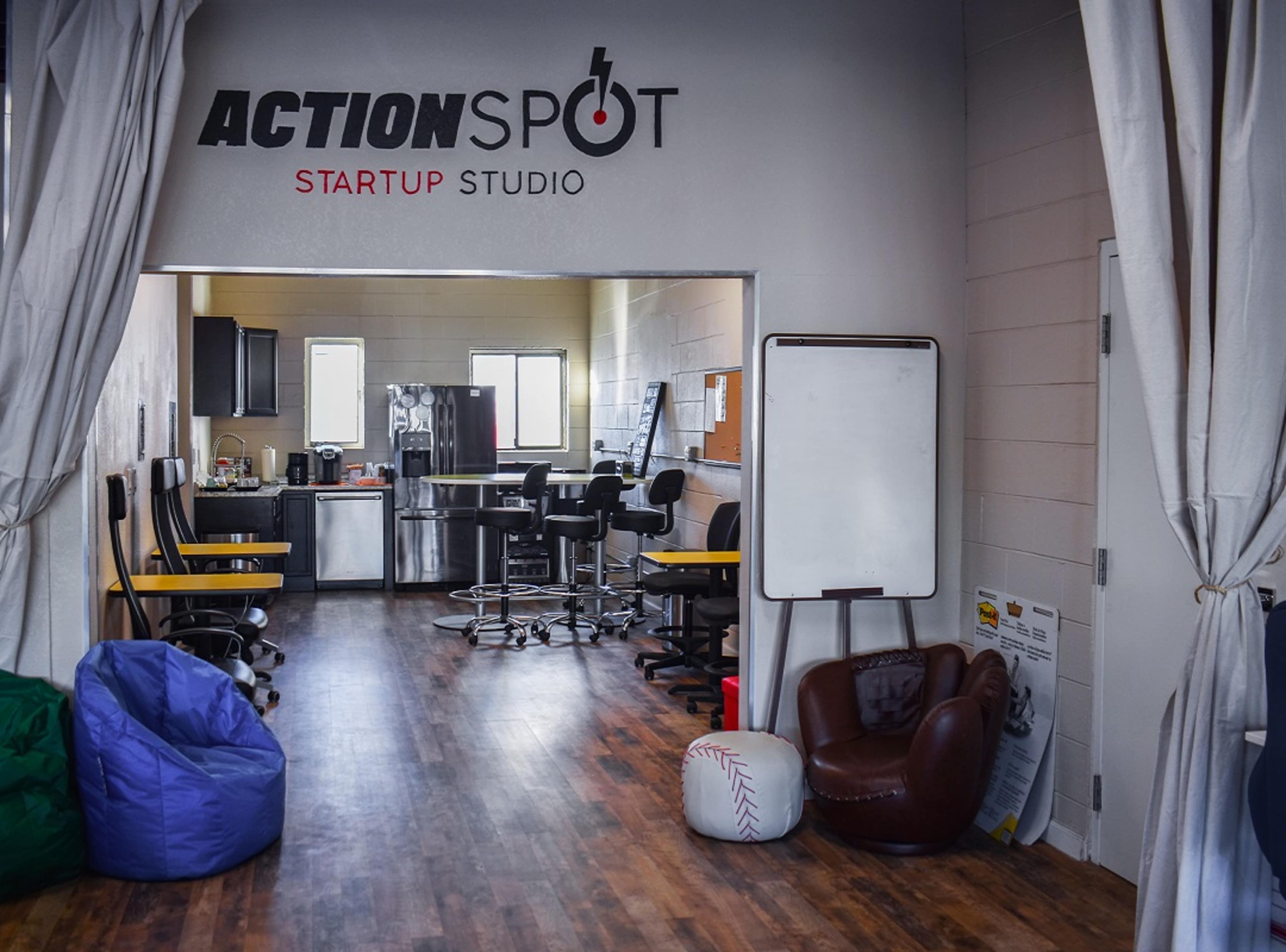 The image shows a modern startup studio with a kitchenette, seating areas, and a large sign that reads "ACTIONSPOT."