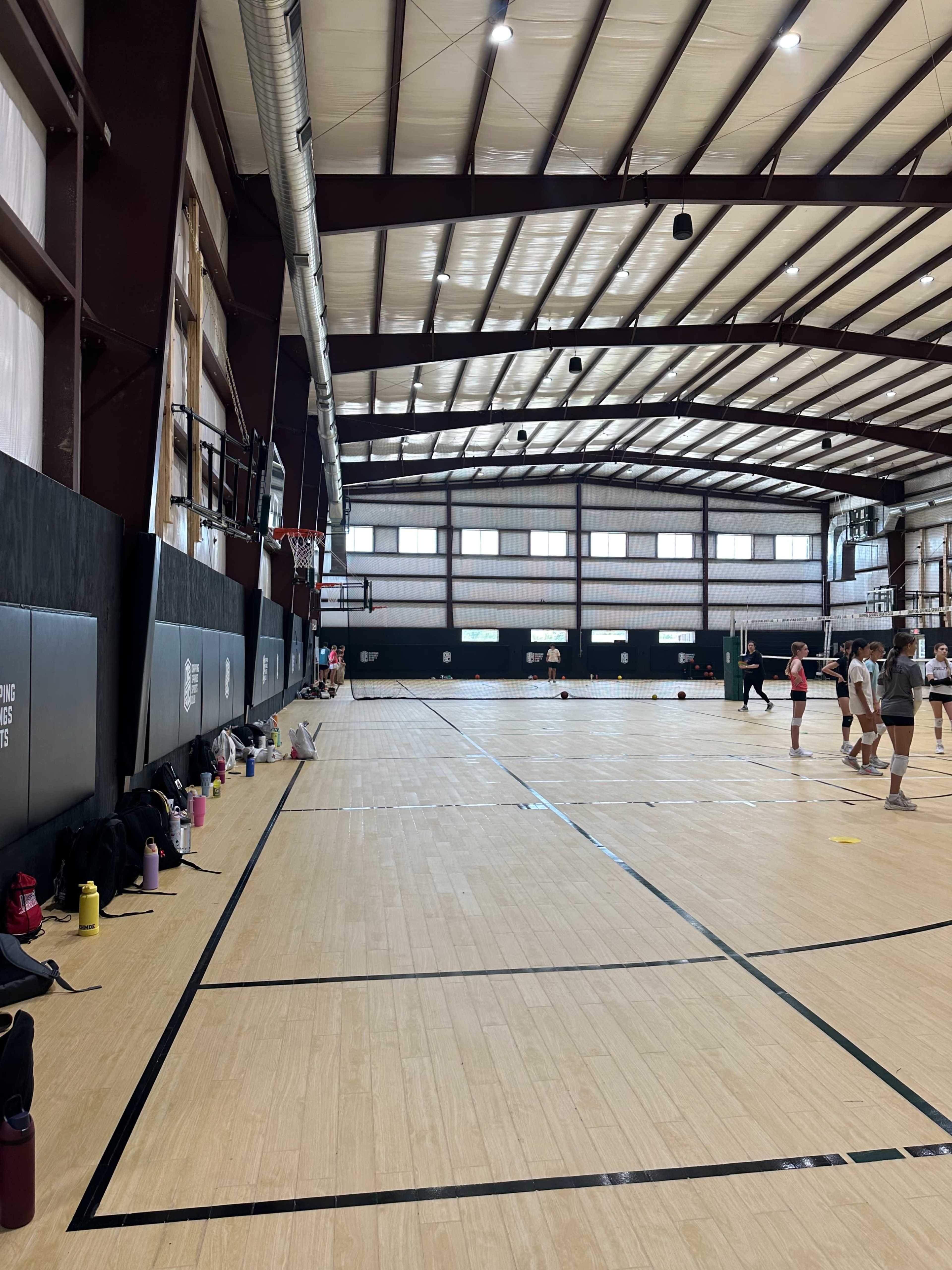 The image shows an indoor sports facility with a wooden floor, basketball hoops, and players engaged in an activity towards the far end.