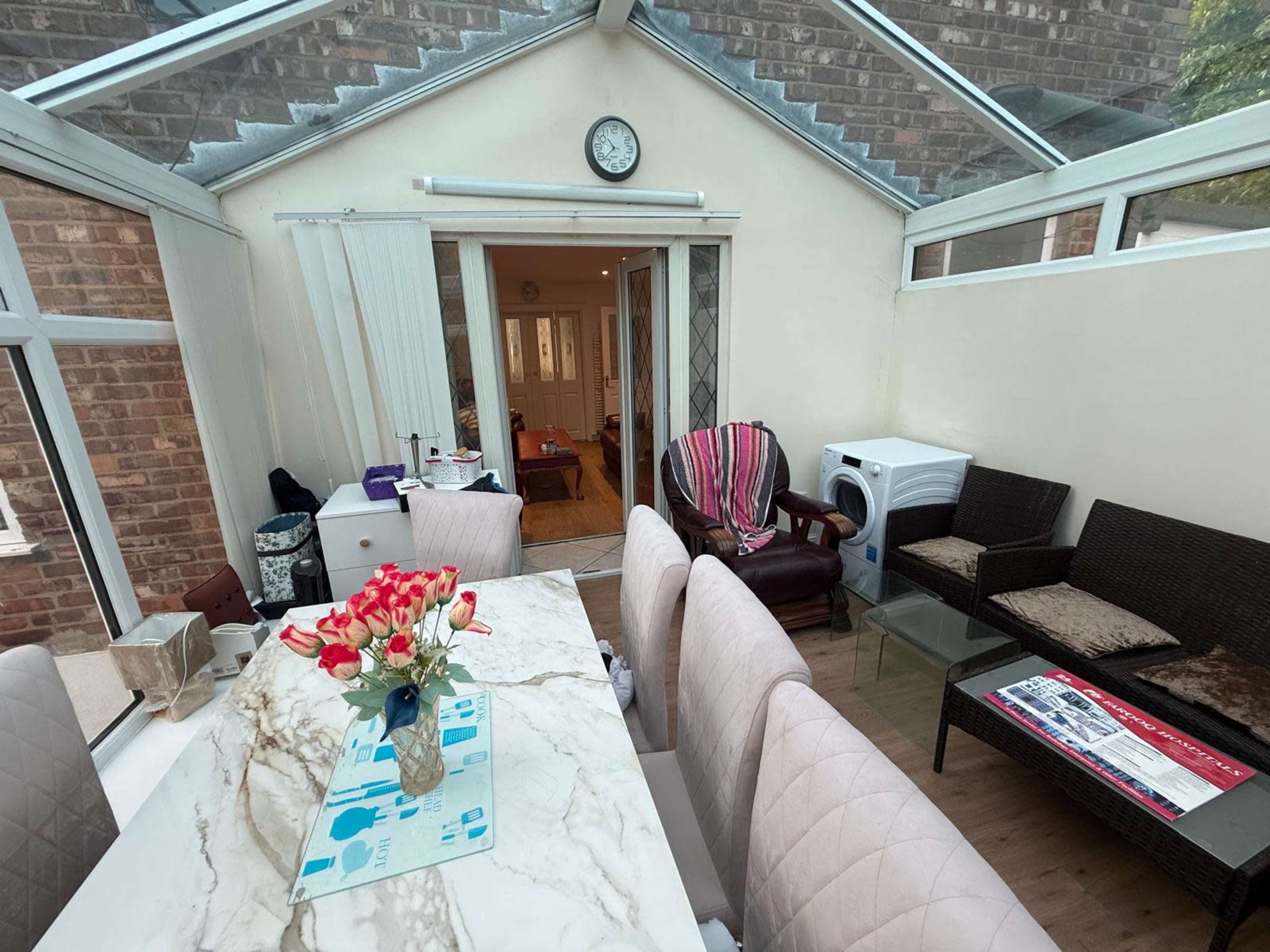The image shows a conservatory with a large table adorned with a vase of flowers, surrounded by chairs, and an open doorway leading to another room.