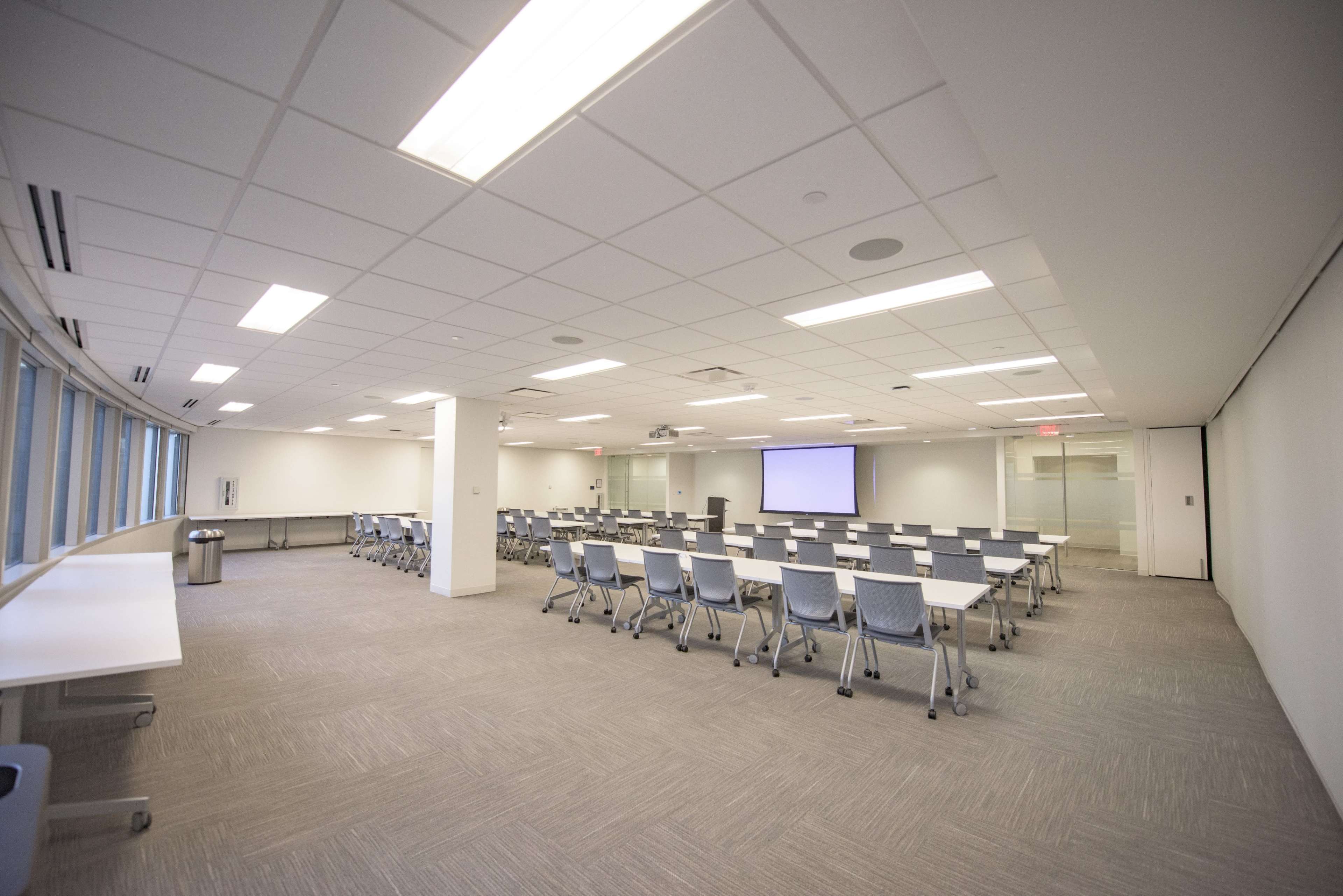 A large, empty conference room with rows of tables and chairs arranged for a meeting, equipped with a projector screen at one end.