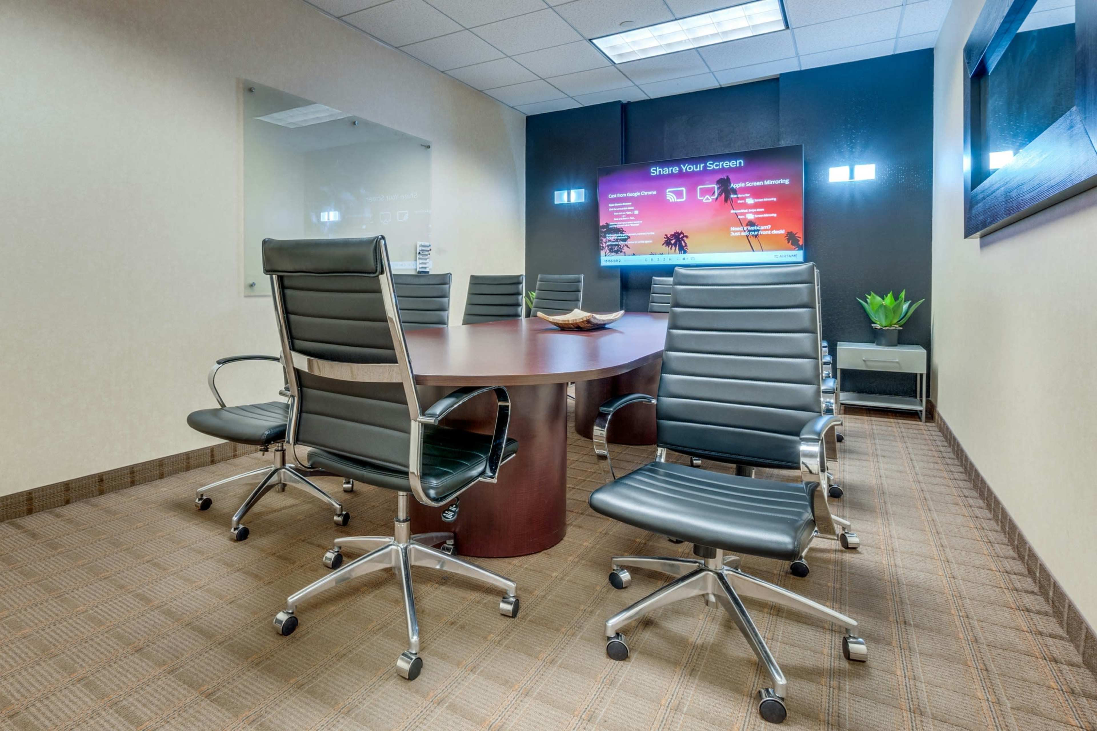 A modern conference room features a round table surrounded by ergonomic chairs and a large screen displaying a "Share Your Screen" message.