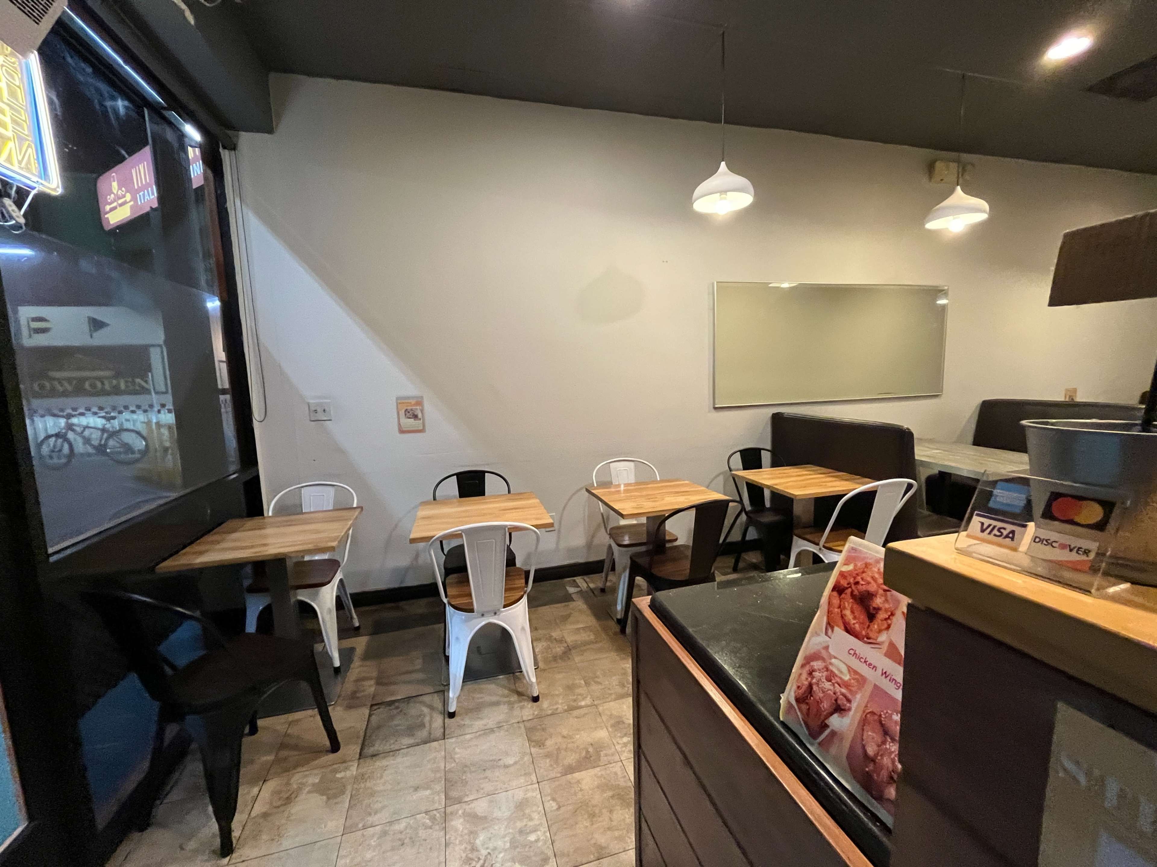 The image shows an empty dining area of a restaurant with several wooden tables and metal chairs arranged on a tiled floor.