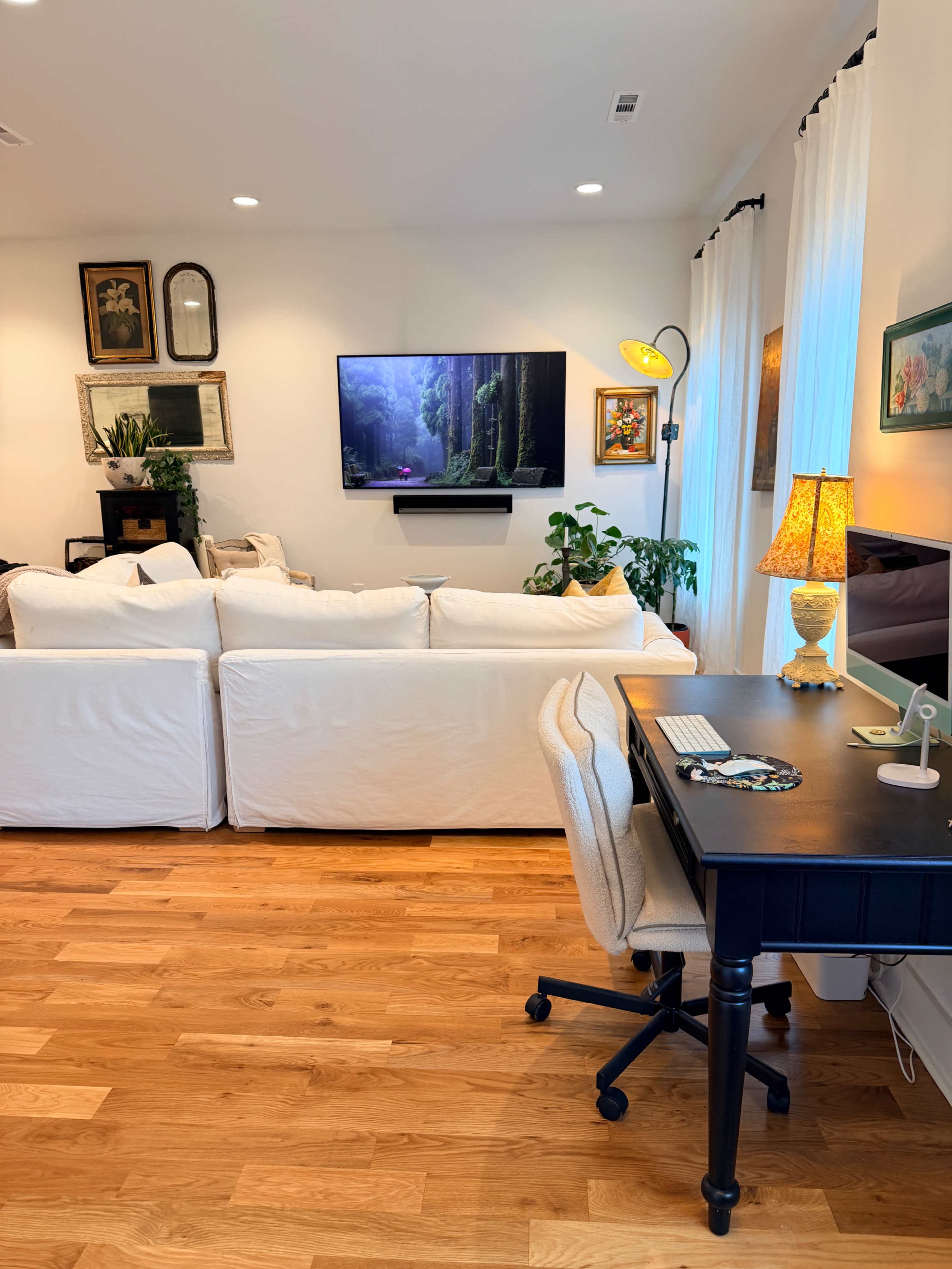 A cozy living room features a white sectional sofa, a wall-mounted TV, houseplants, and a desk with a computer.