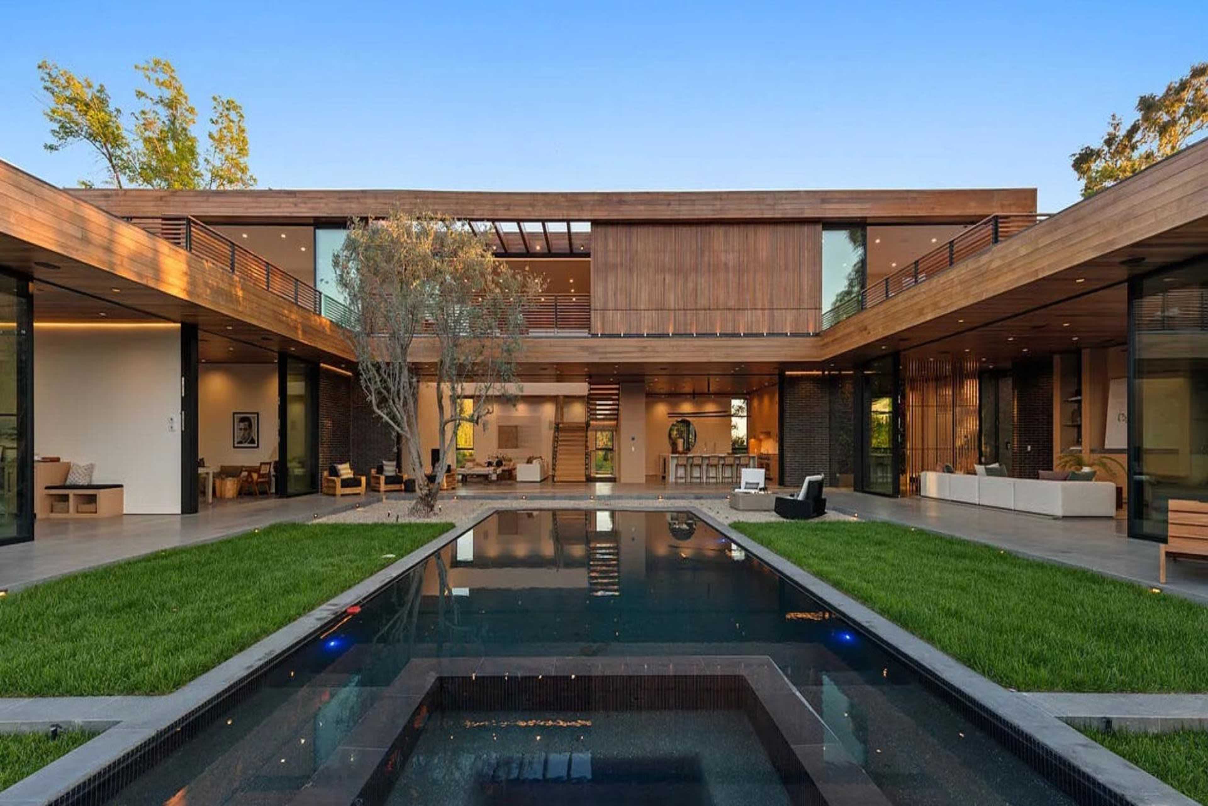 The image shows a modern two-story house with large glass windows, wooden accents, and an outdoor pool surrounded by grass.