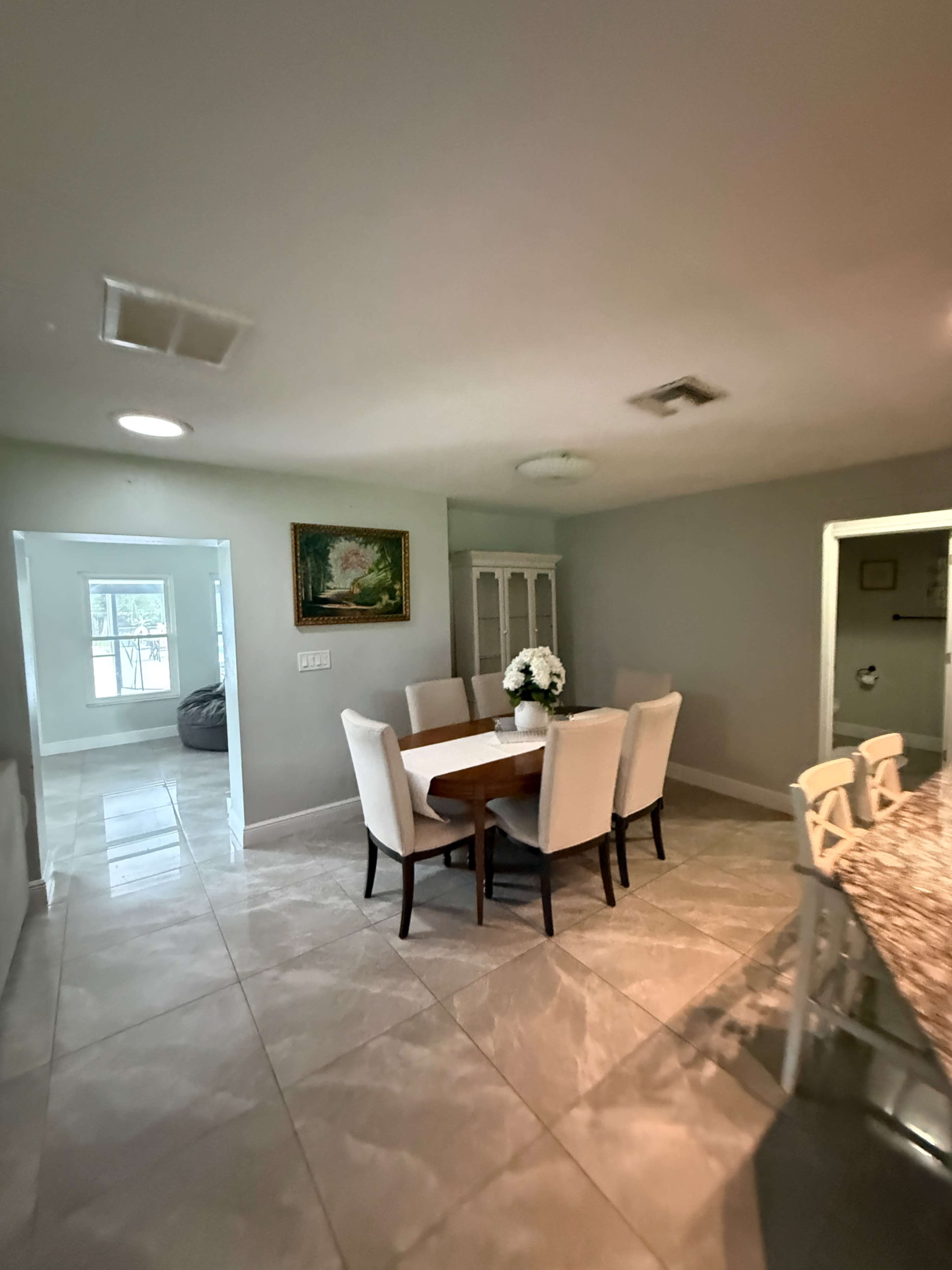 The image shows a dining area with a wooden table surrounded by six upholstered chairs, adjacent to a kitchen and a secondary room featuring a large window.