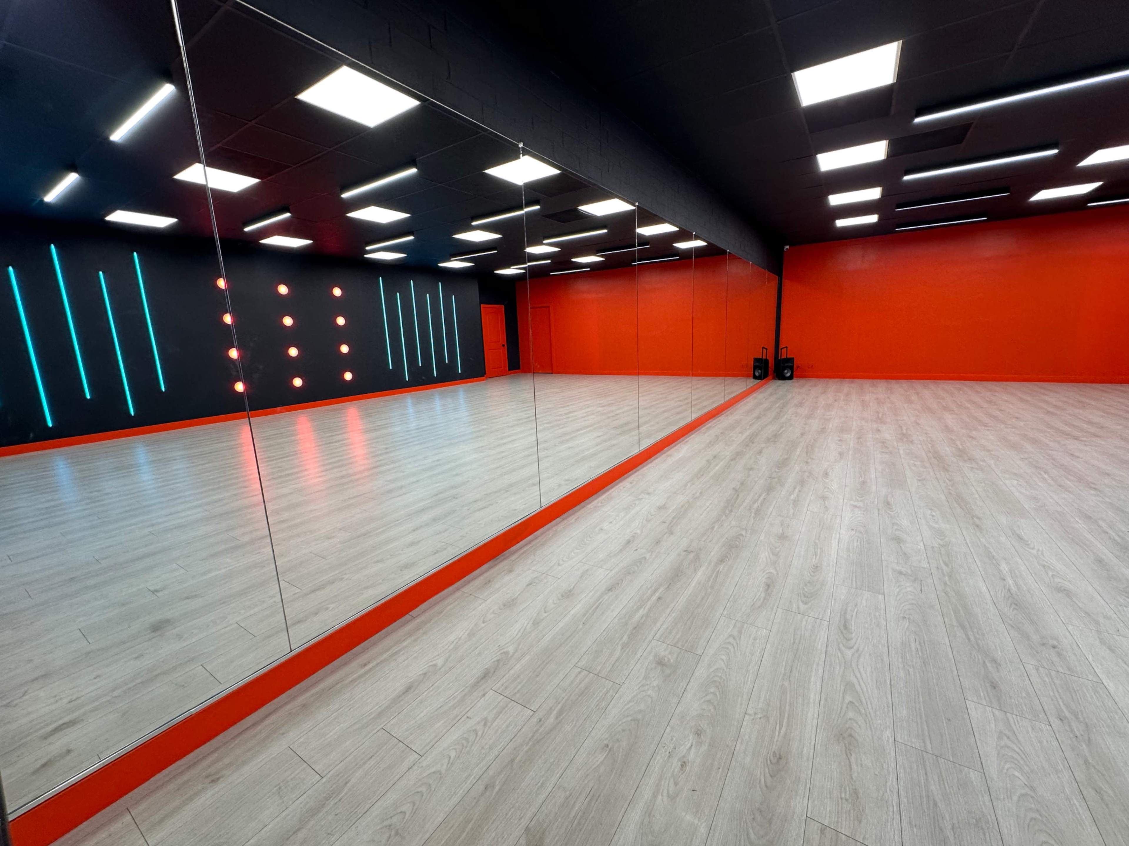 A spacious dance studio featuring a large mirrored wall, wooden flooring, and vibrant red and black wall accents with LED lighting.