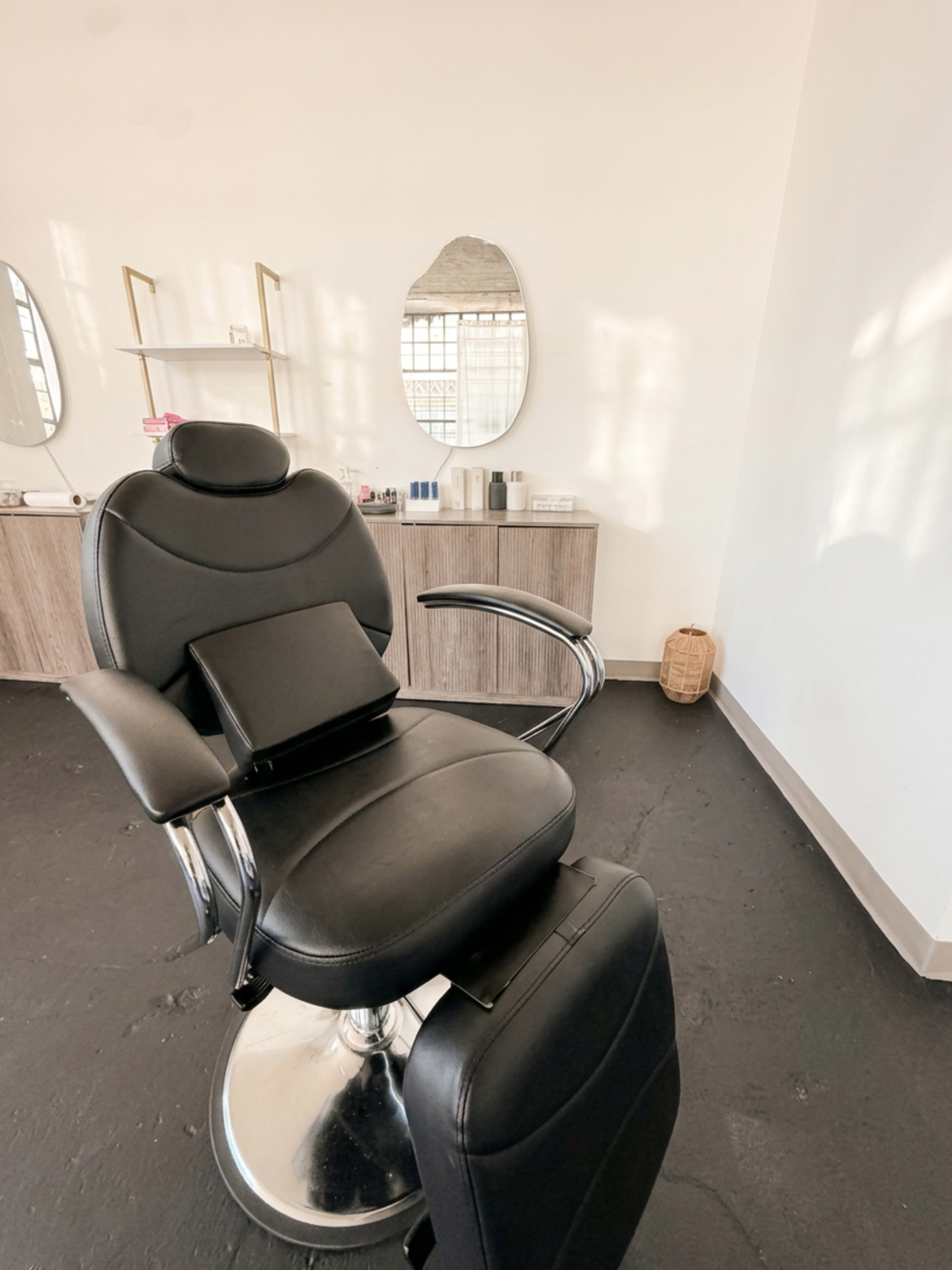 The image shows a black salon chair in a brightly lit room with a mirror and a wooden cabinet in the background.
