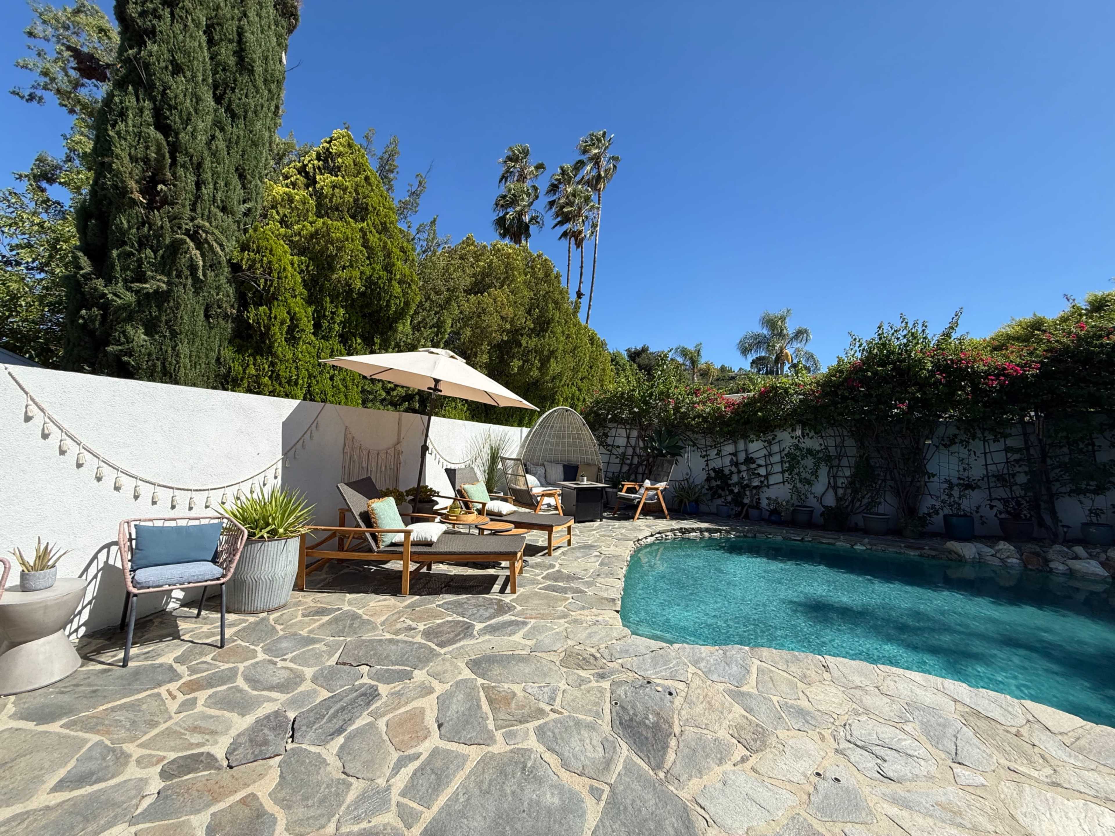 The image shows a landscaped backyard with a swimming pool, stone patio, lounge chairs, and an umbrella surrounded by greenery and palm trees.