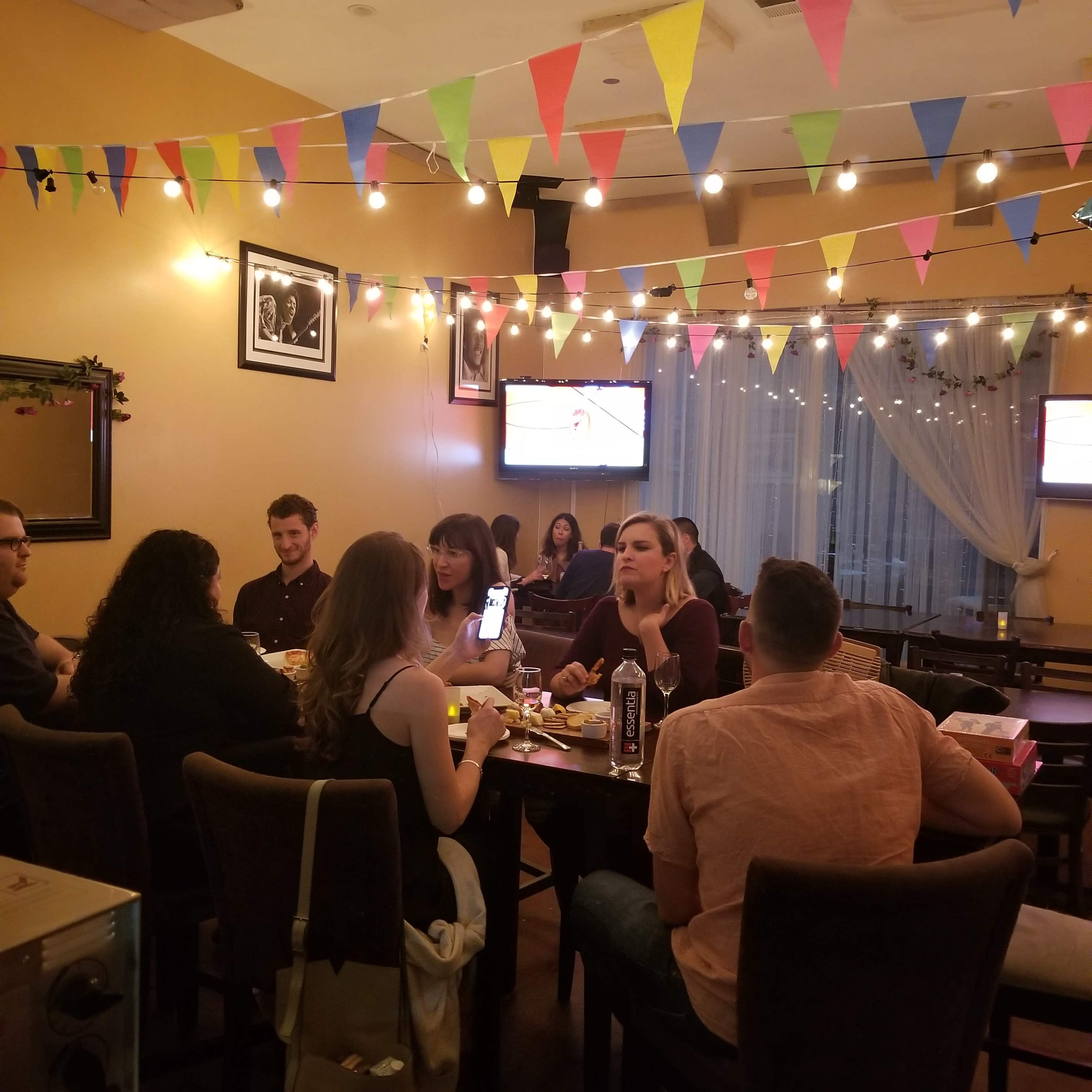 A group of people gathers around tables in a brightly decorated restaurant with string lights and colorful bunting, while two televisions display content in the background.
