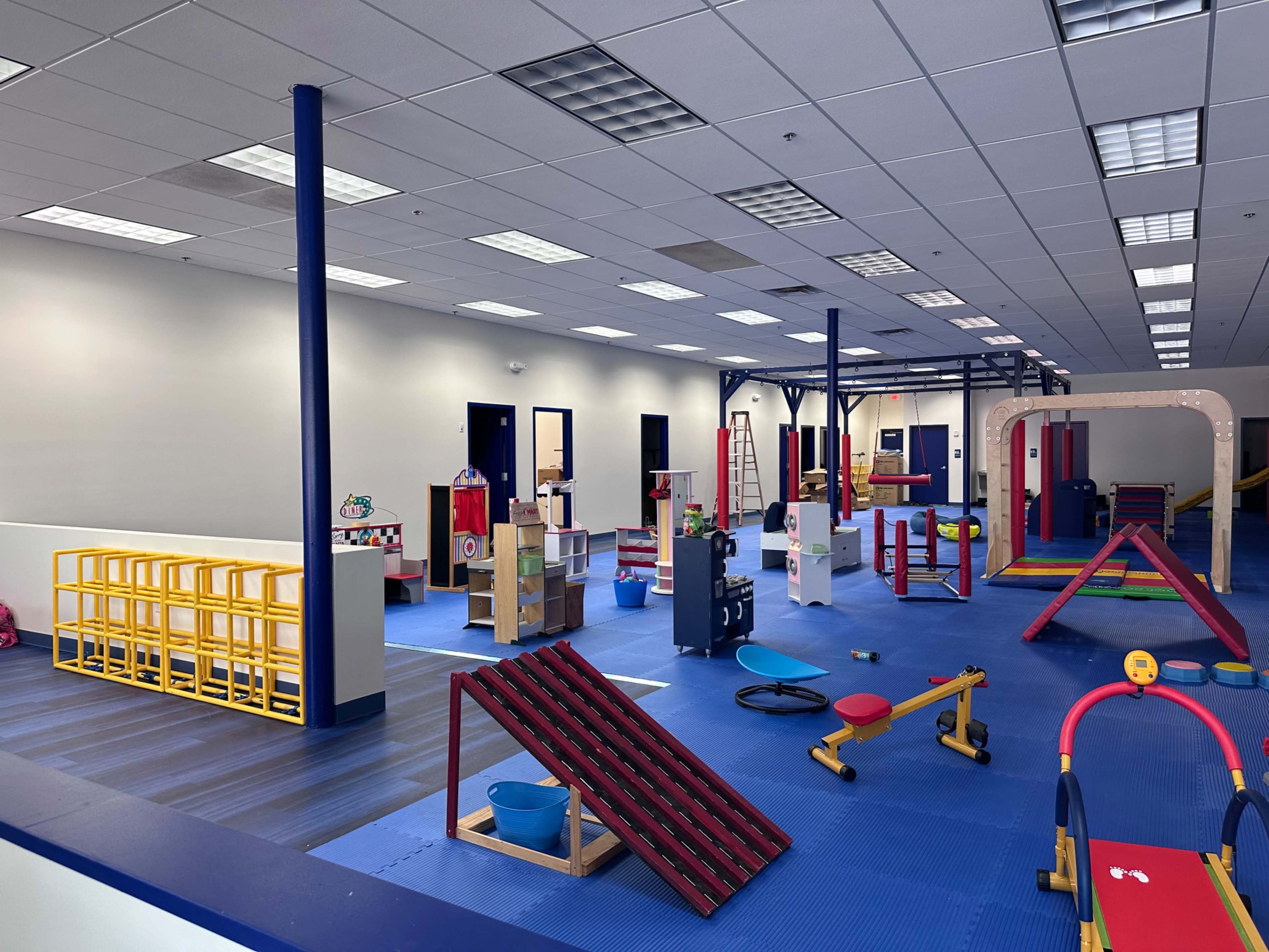 The image shows a brightly lit playroom filled with various play structures, toys, and play equipment arranged on a blue floor.