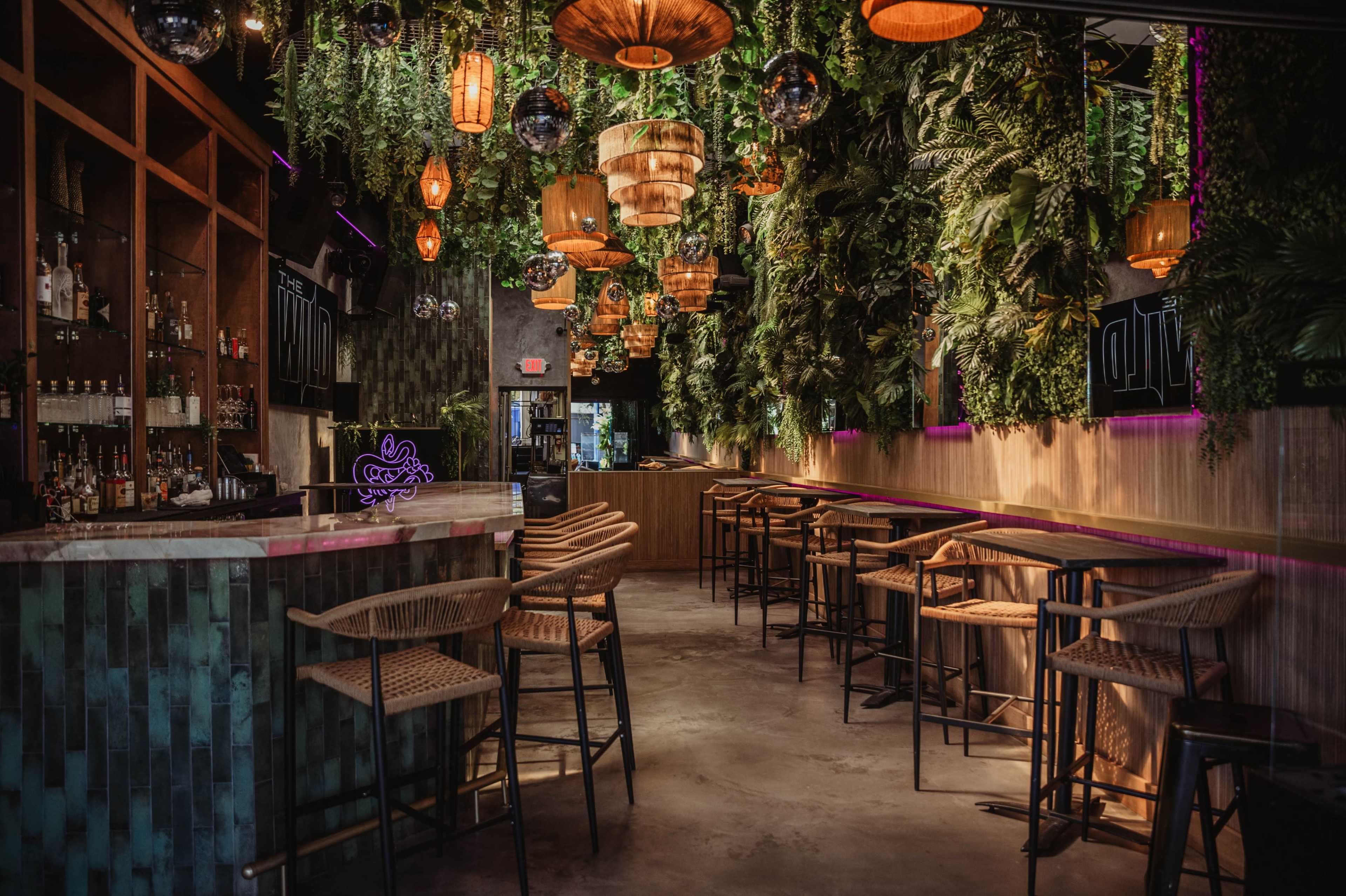 The image depicts a modern bar interior with a lush backdrop of hanging plants, pendant lights, and a sleek counter with high chairs.