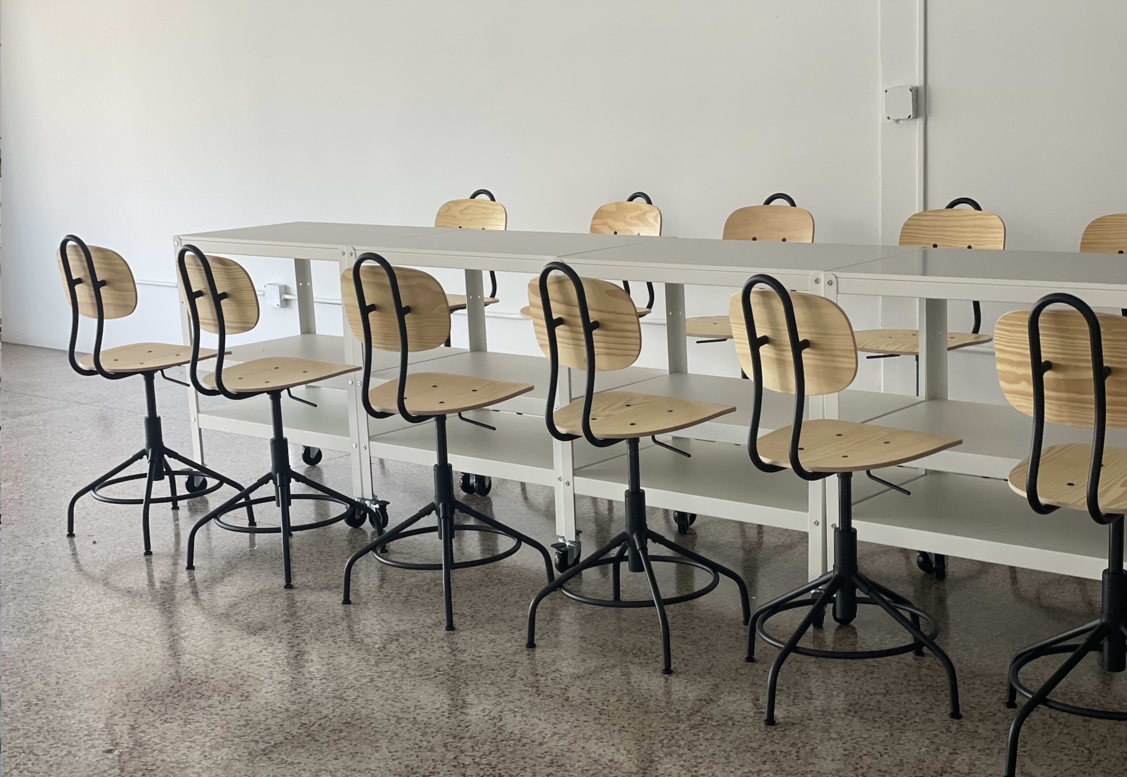 A row of wooden stools with metal bases is positioned around a long white table in a minimalist room.