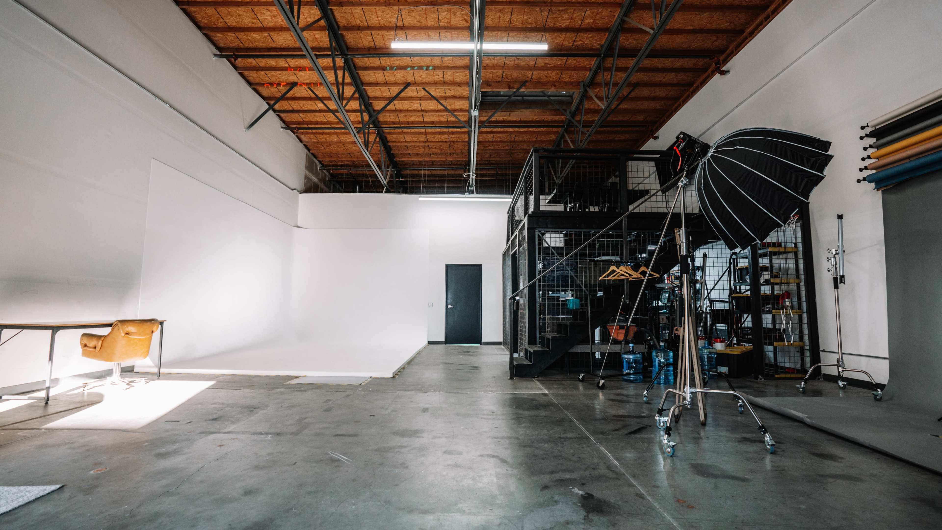 A large, empty studio space with a white backdrop, a metal staircase leading to an upper storage area, and photography equipment arranged on one side.
