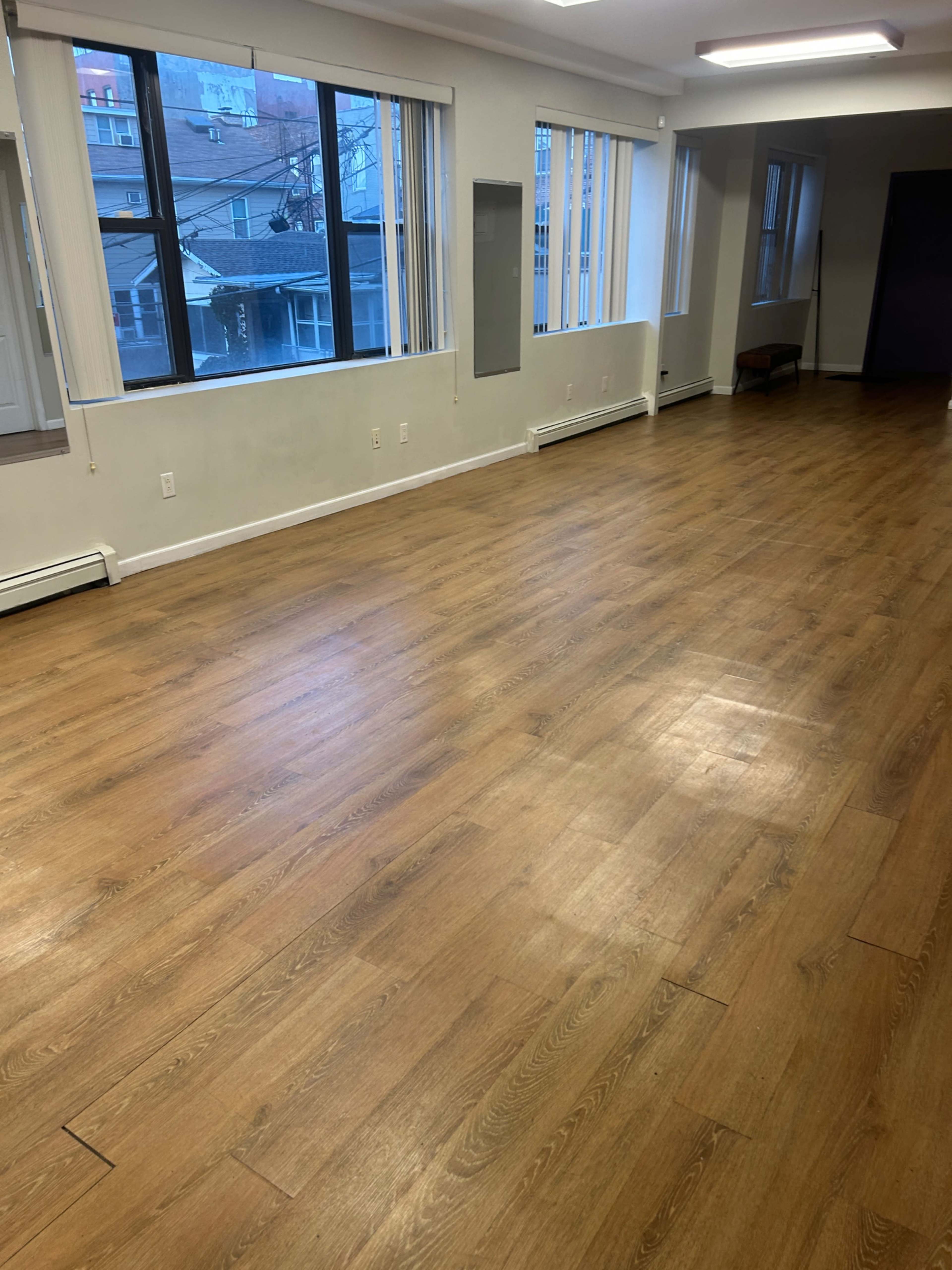 The image shows a spacious room with wooden flooring, large windows, and minimal furniture.