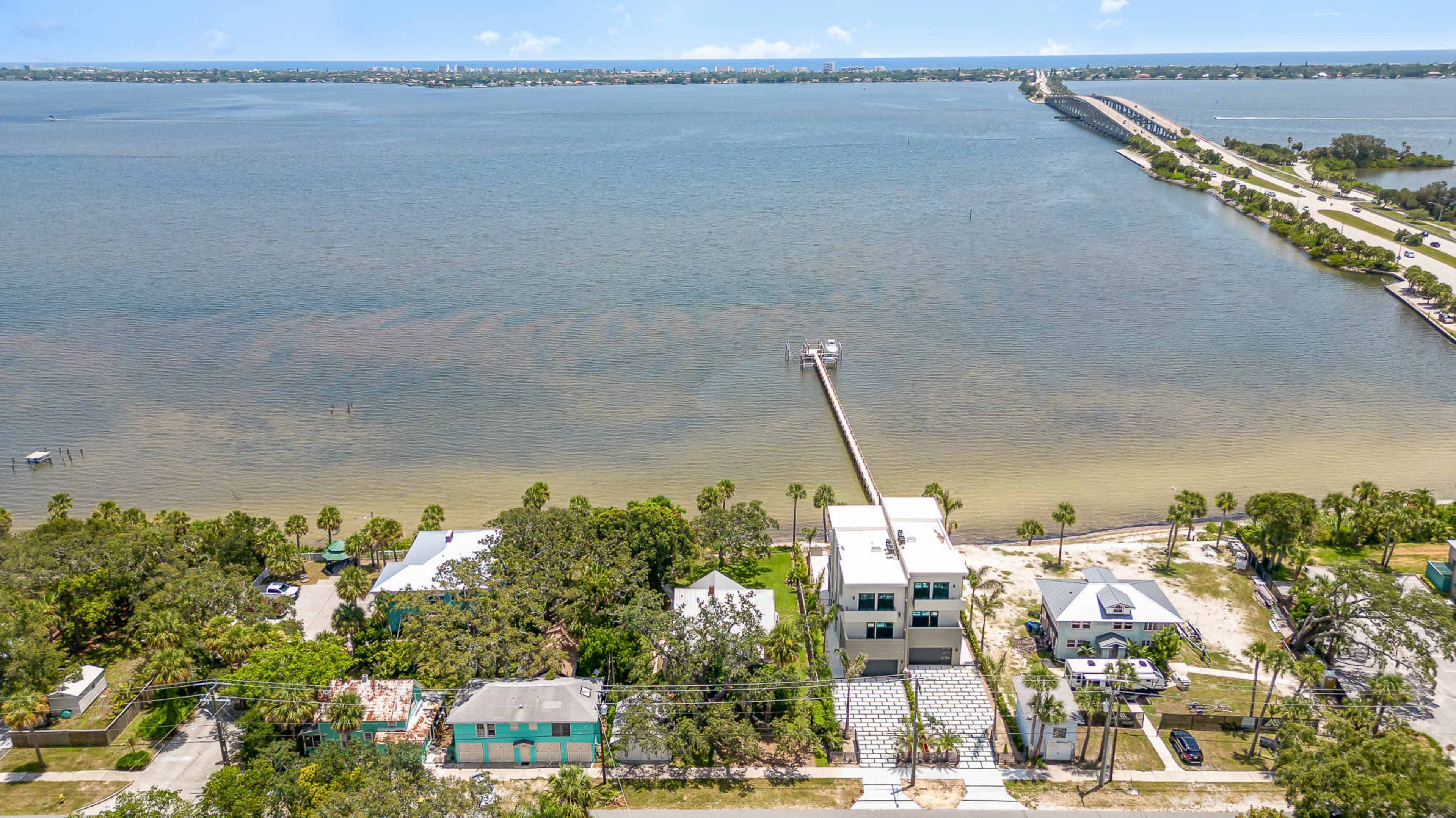 An aerial view of a waterfront area featuring various houses, a long pier extending into the water, and a bridge in the distance.