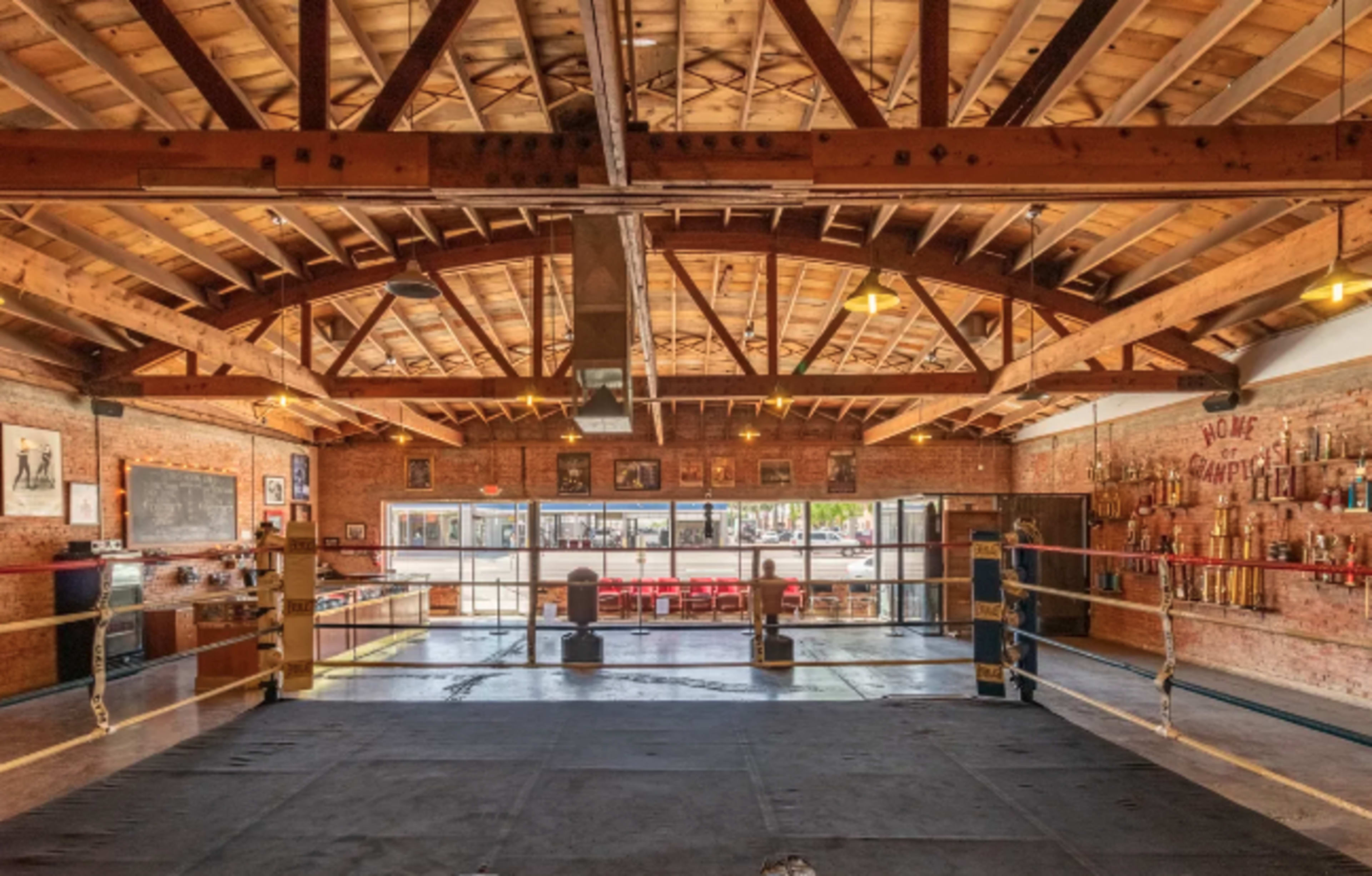 The image shows a boxing gym with a central ring surrounded by wooden beams and brick walls, adorned with trophies and photographs.