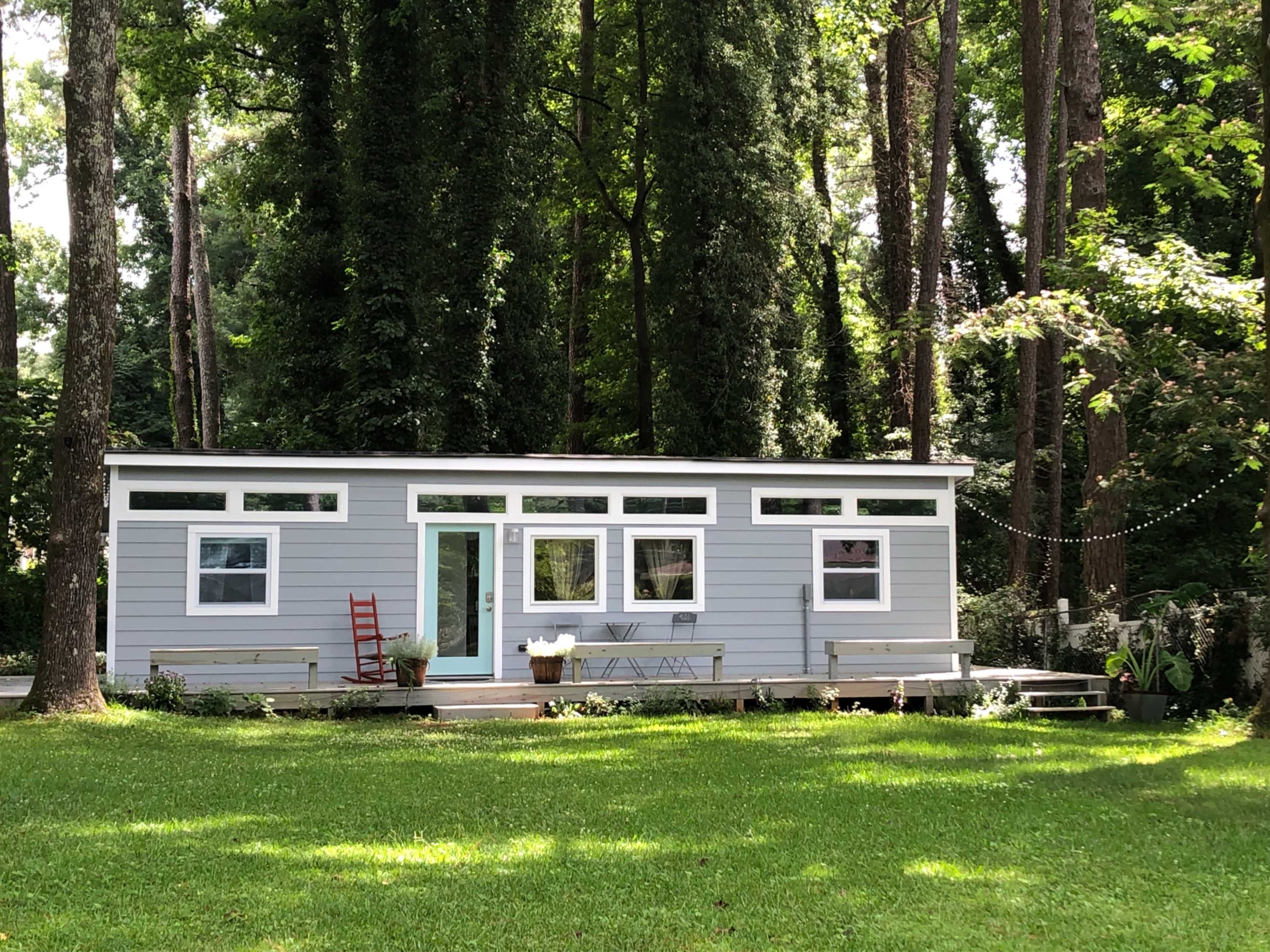 A modern, single-story cabin with large windows is set on a green lawn surrounded by tall trees.