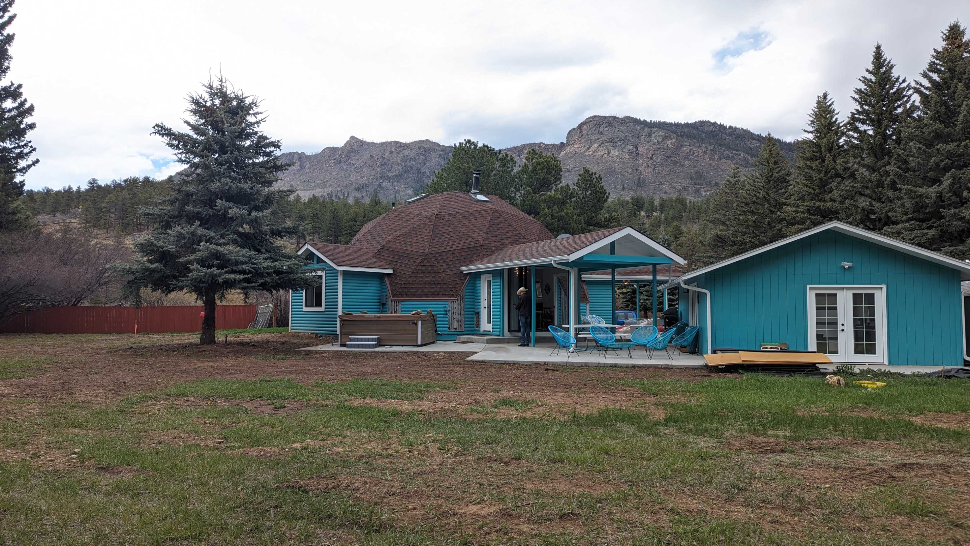 A turquoise domed house with a sloped roof and a small adjacent building is set against a backdrop of mountains and trees.