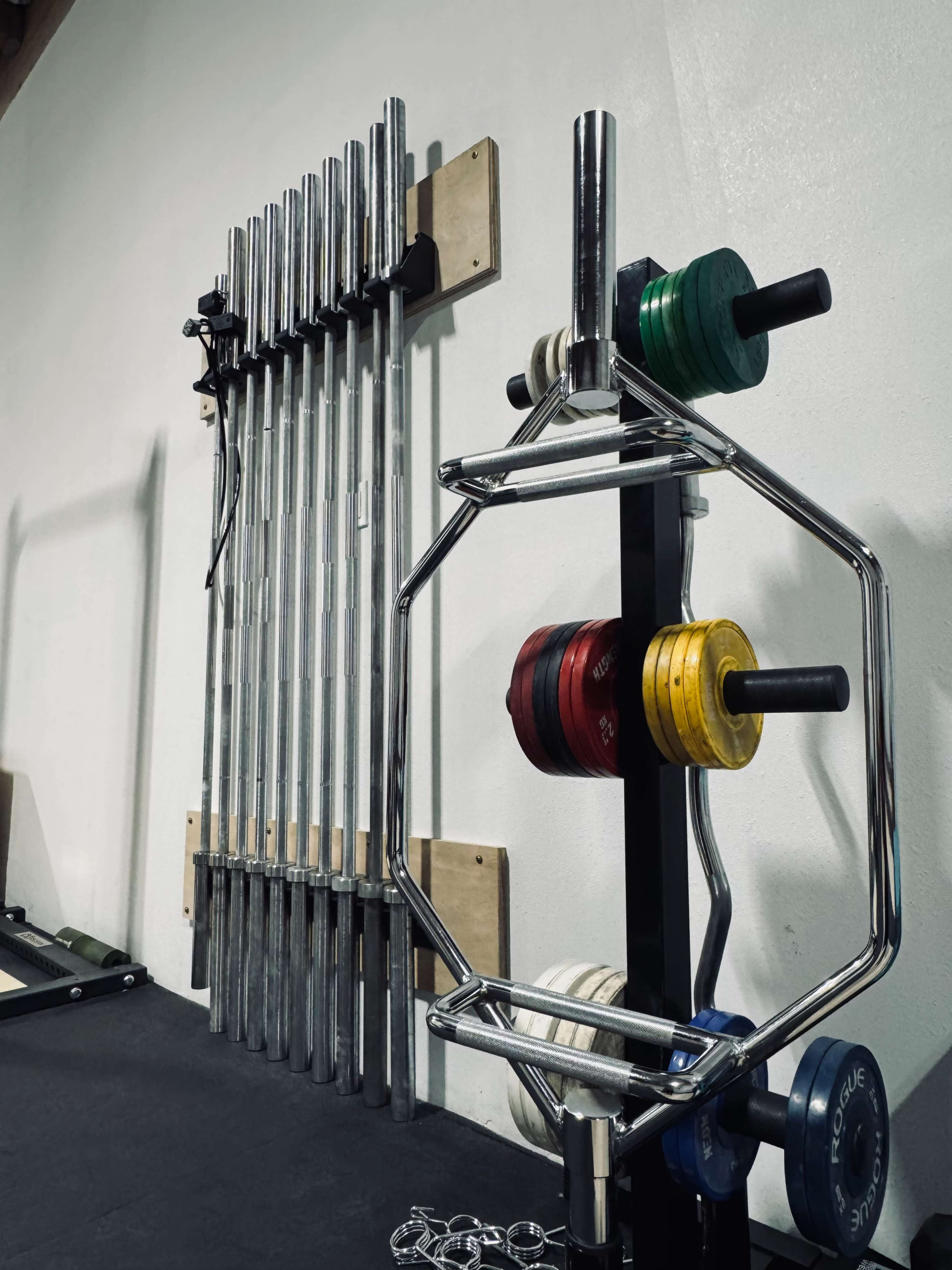The image shows a gym interior with a vertical rack holding various weight plates and a set of long barbells mounted on the wall.