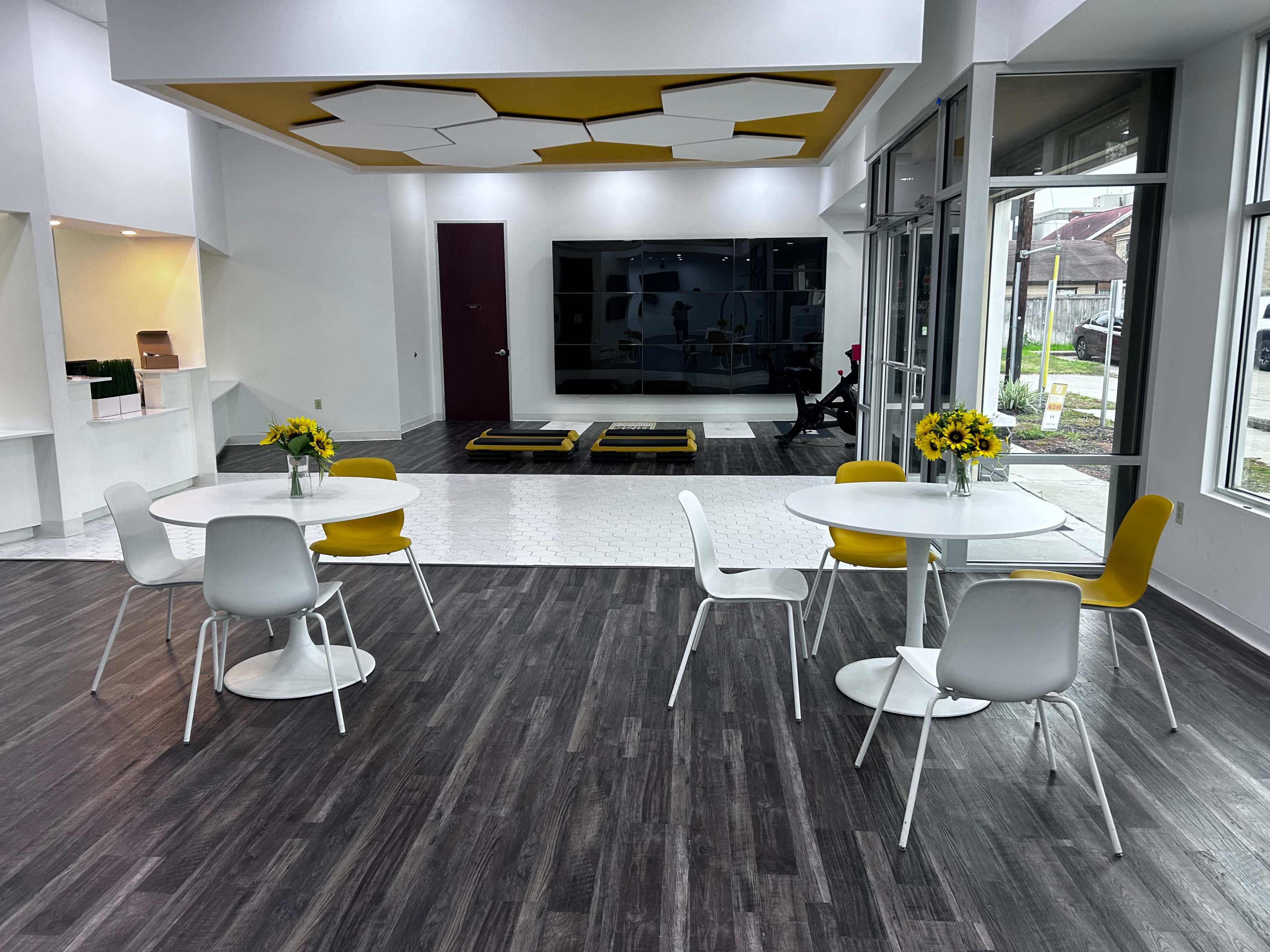 The image shows a modern, open space featuring two round tables with yellow chairs, a large wall-mounted screen, and a section with a patterned white floor.