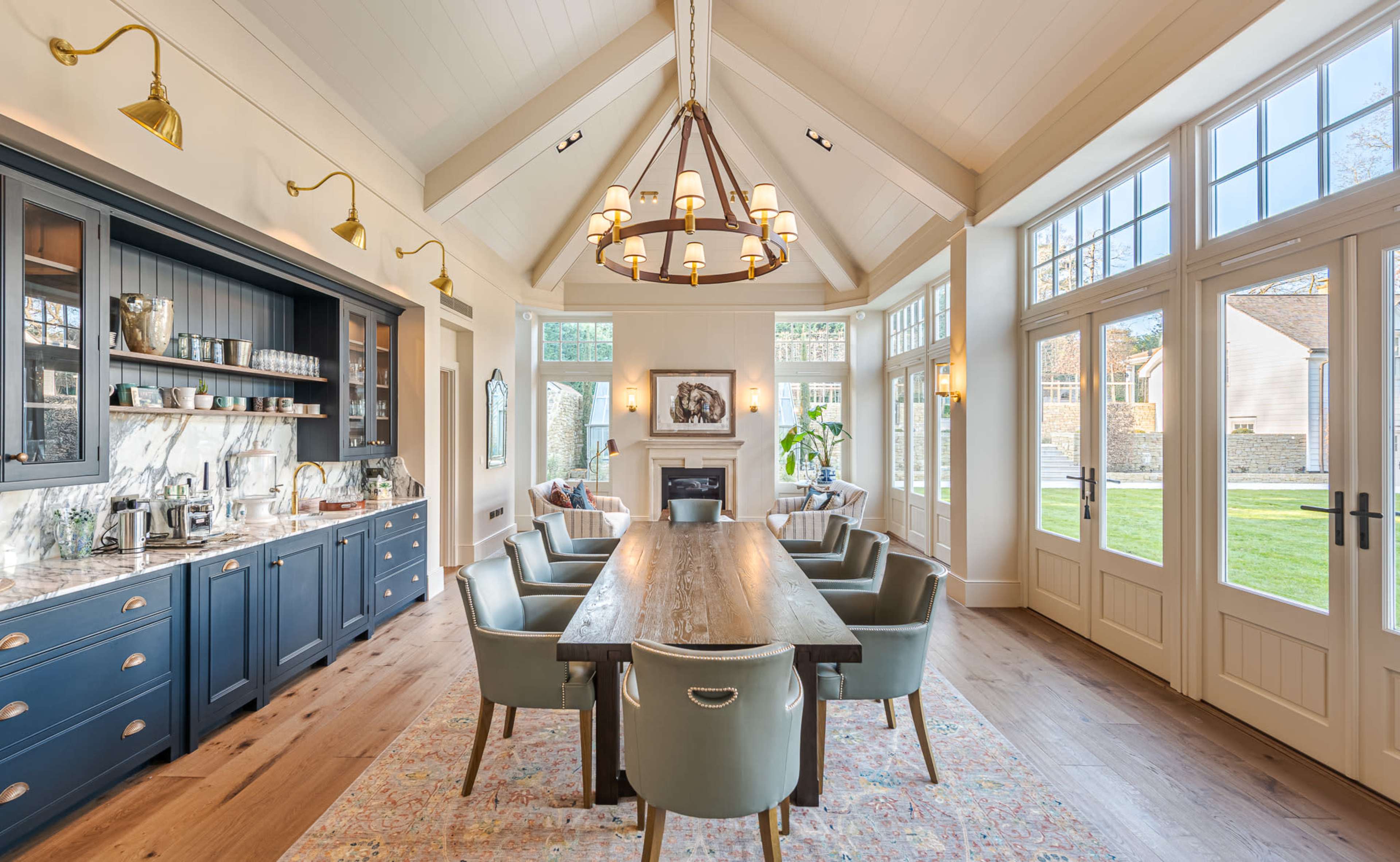 A spacious dining area features a long wooden table surrounded by upholstered chairs, large windows allowing natural light, and a decorative chandelier overhead.