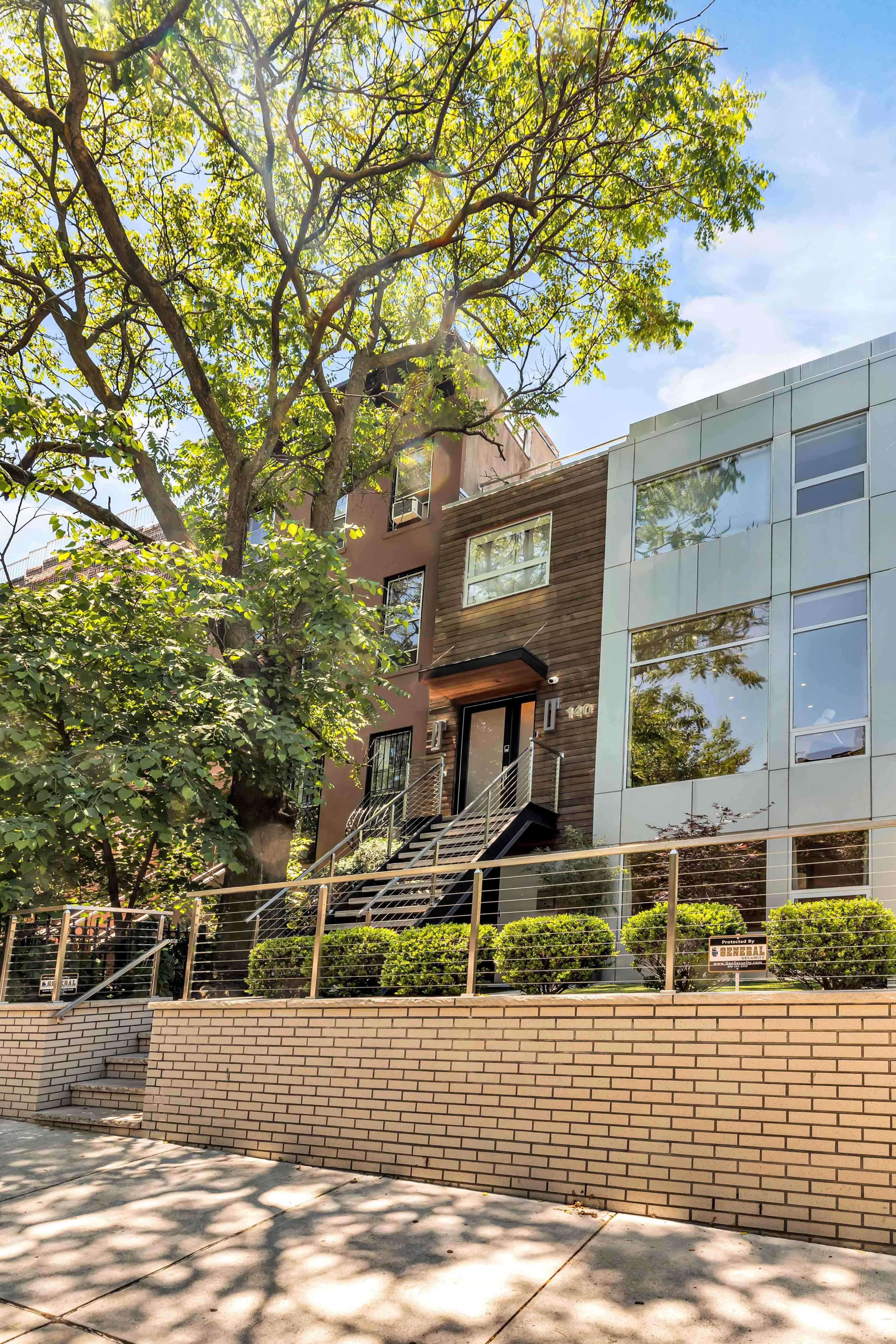 A modern multi-story building with a blend of brown and glass facades, situated beside a large tree and surrounded by well-manicured greenery.