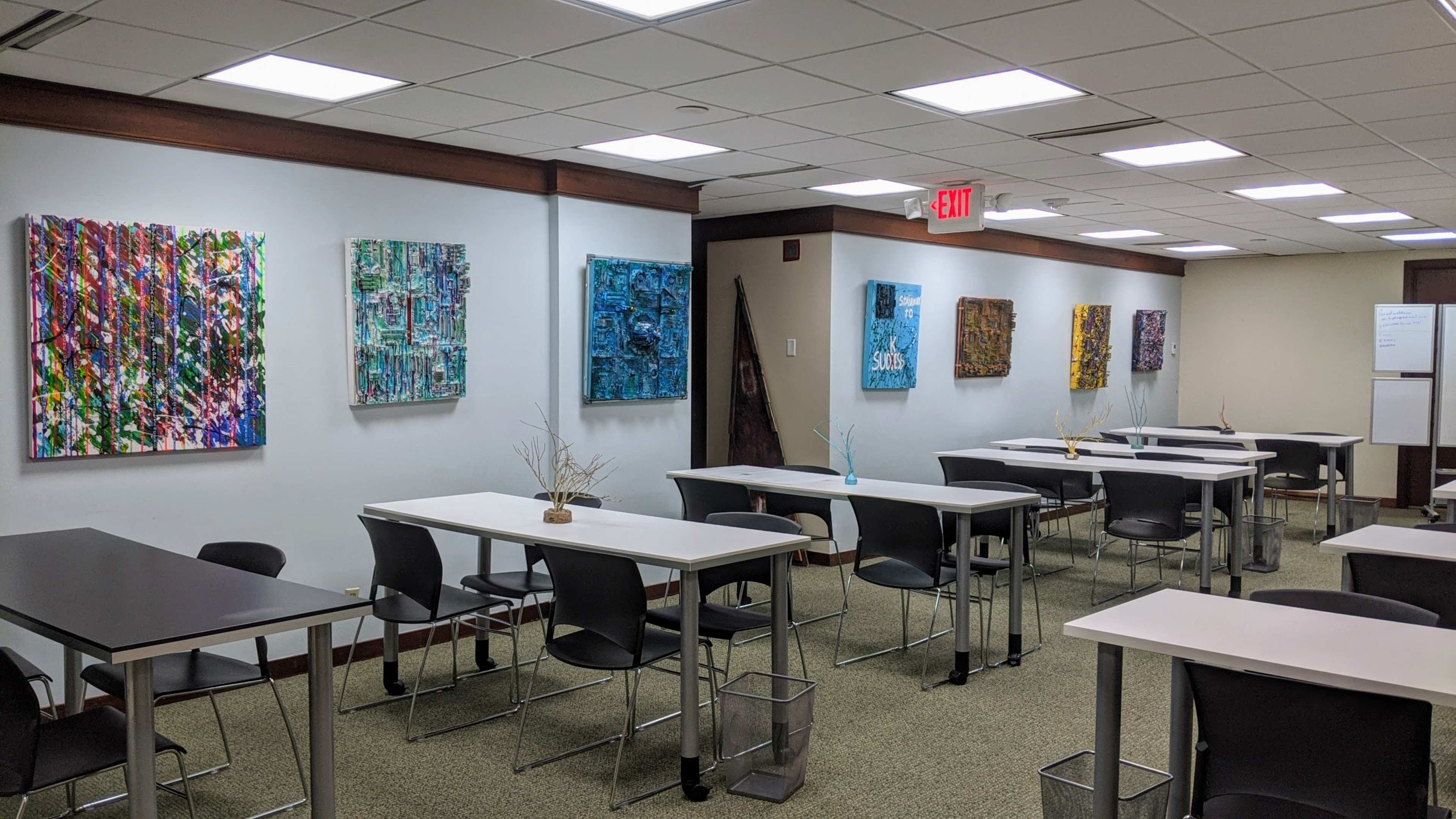 The image shows a modern meeting room with multiple tables and chairs arranged around, featuring colorful artwork hanging on the walls.