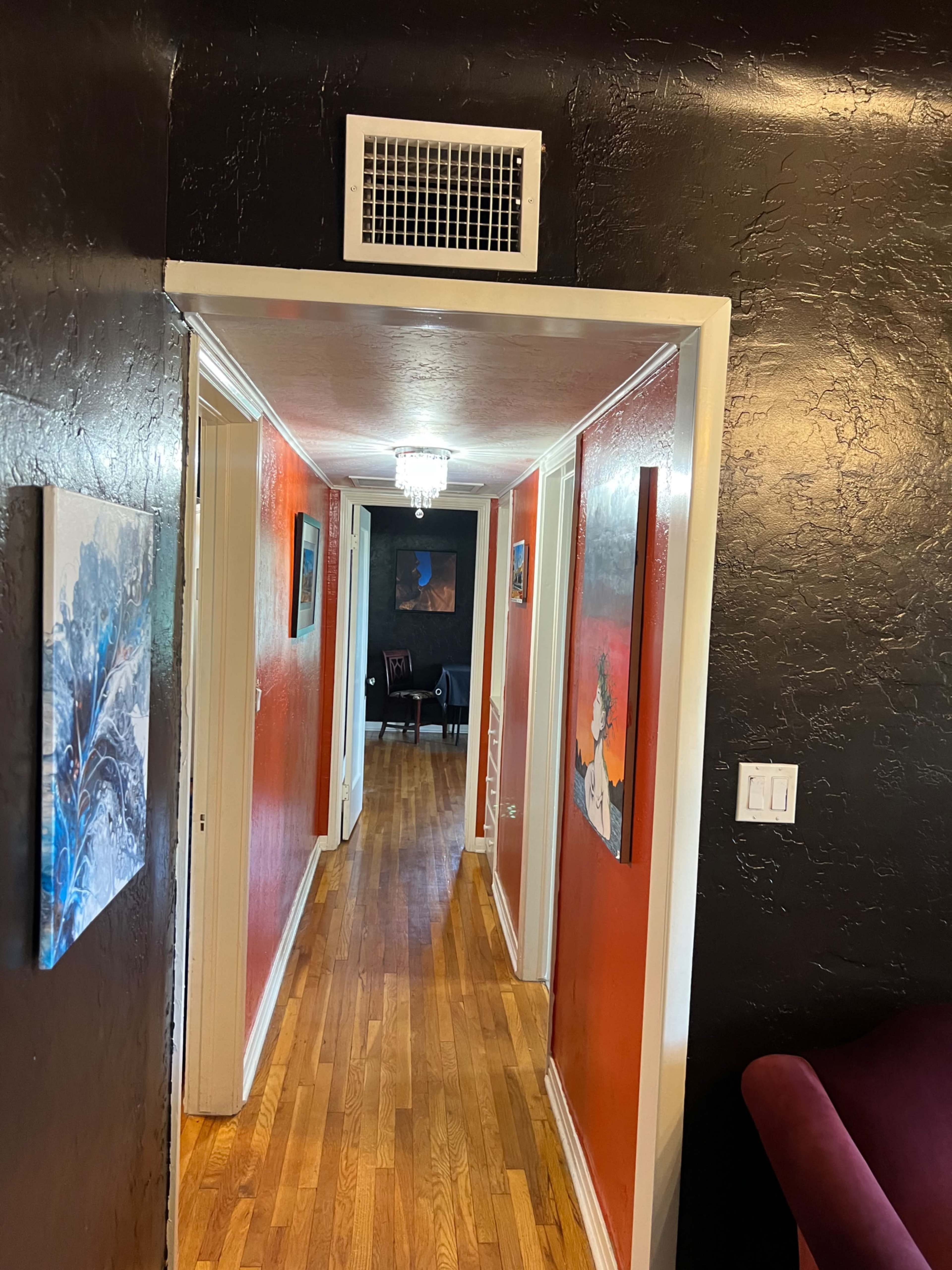 A hallway with orange walls, hardwood flooring, and several pieces of artwork hanging on the walls, leading to a distant room with a chair.