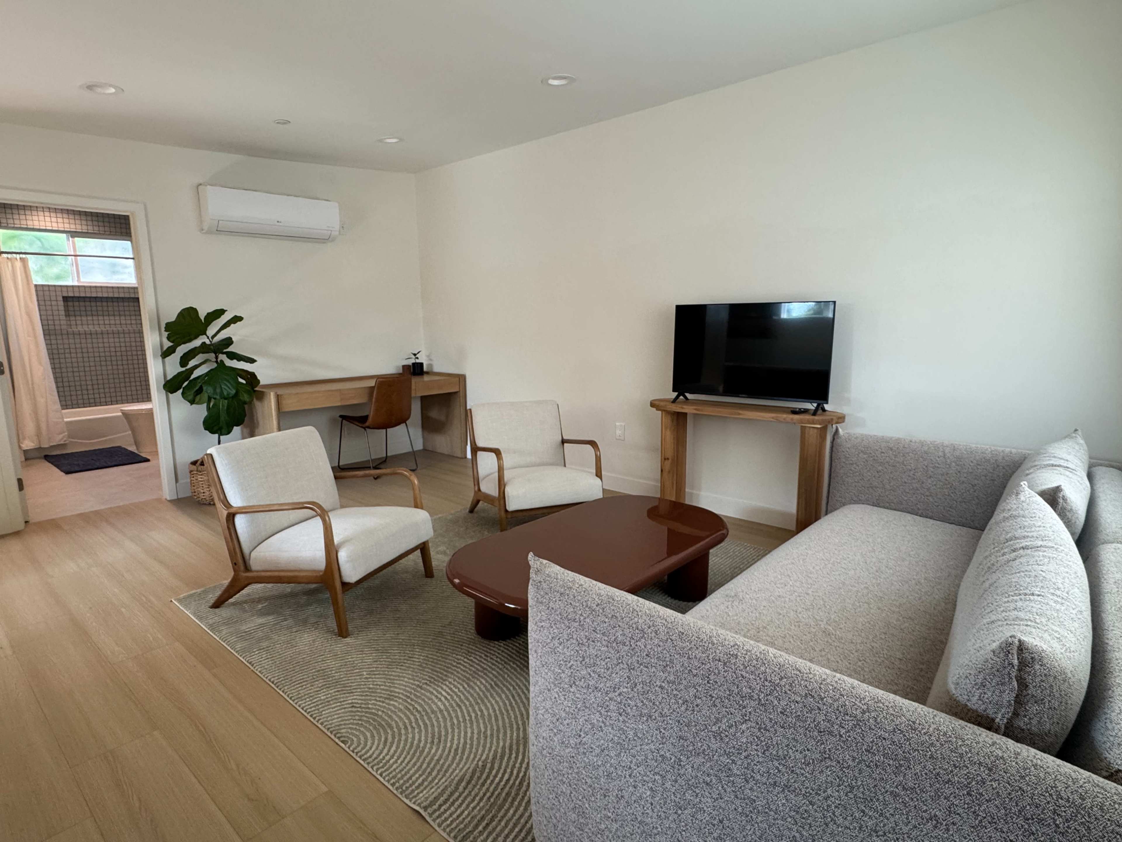 The image shows a modern living room with a gray sofa, two wooden chairs, a coffee table, a television, and a workspace with a desk and chair.