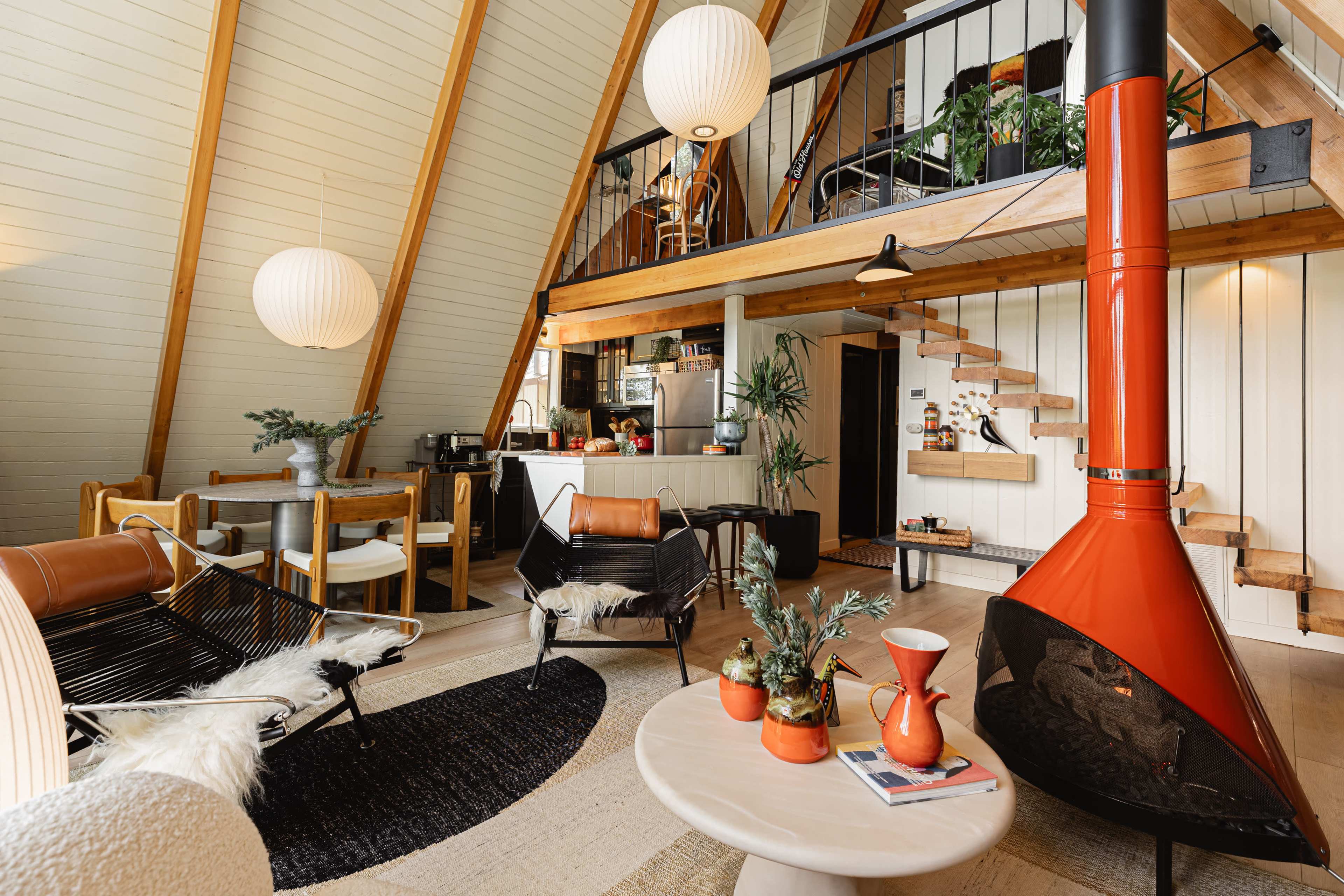 The image shows a modern, loft-style living area featuring a triangular wooden ceiling, a red fireplace, and a mix of seating arrangements, including a round table and a black rug.