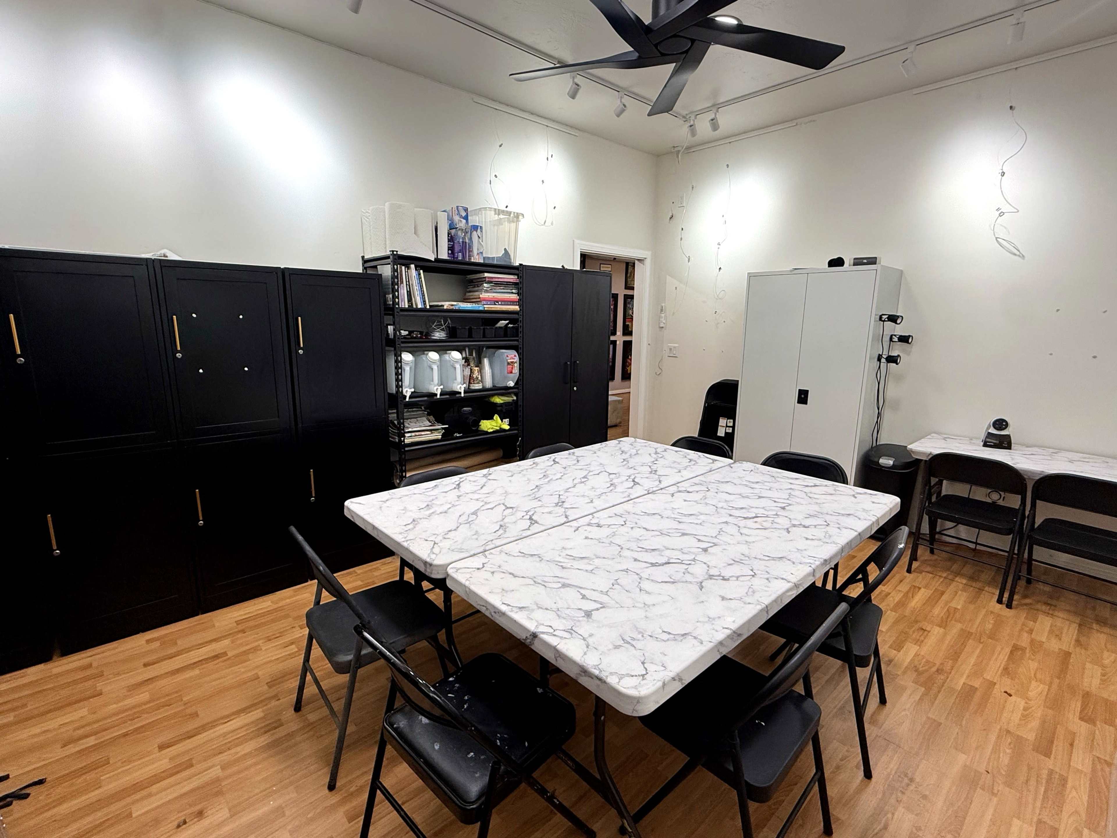 The image shows a room with two tables covered in marble-patterned plastic, surrounded by black folding chairs, and a backdrop of dark storage cabinets and shelves.
