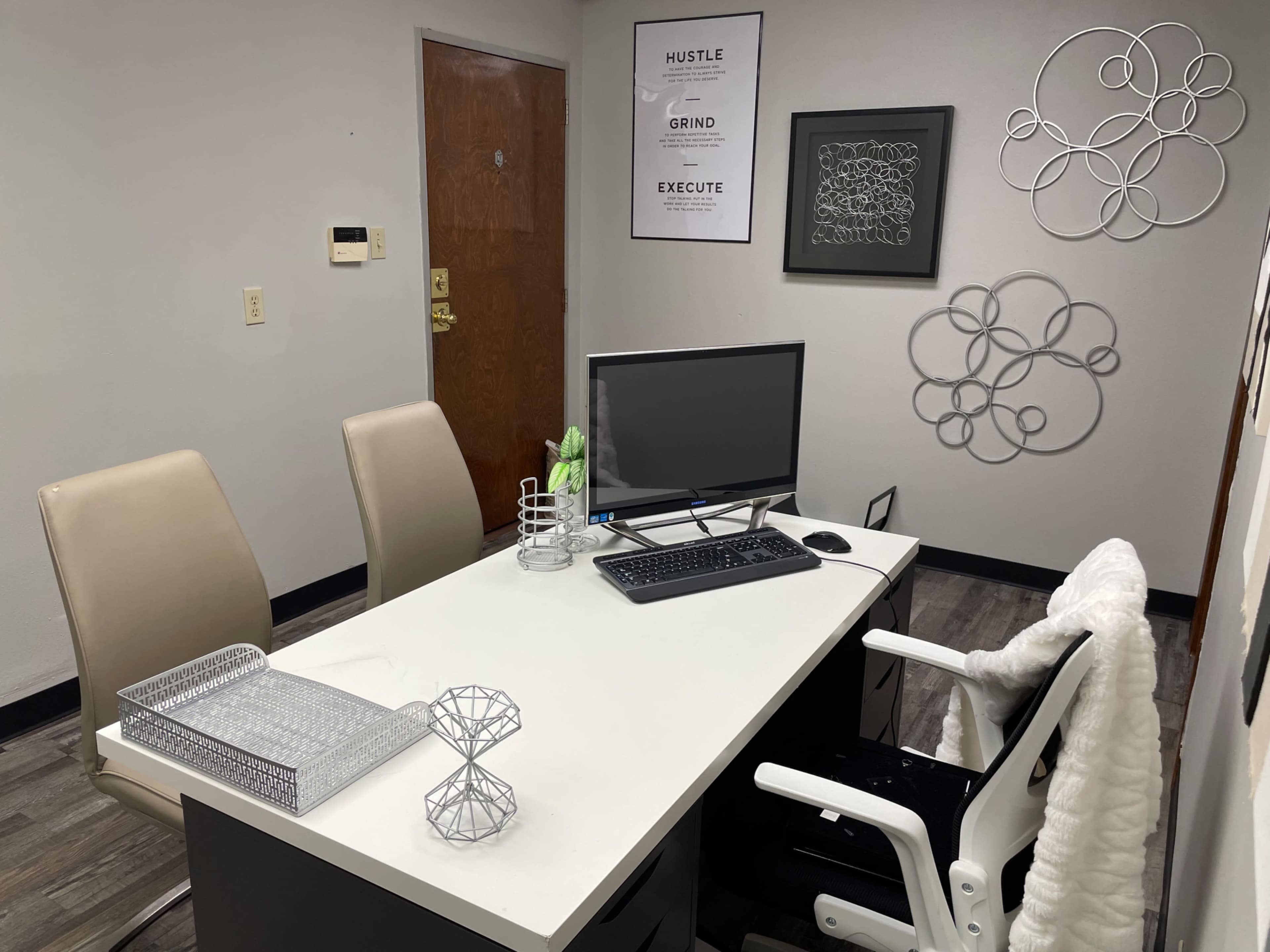 The image shows a small office with a desk, a computer, two chairs, and wall decorations.