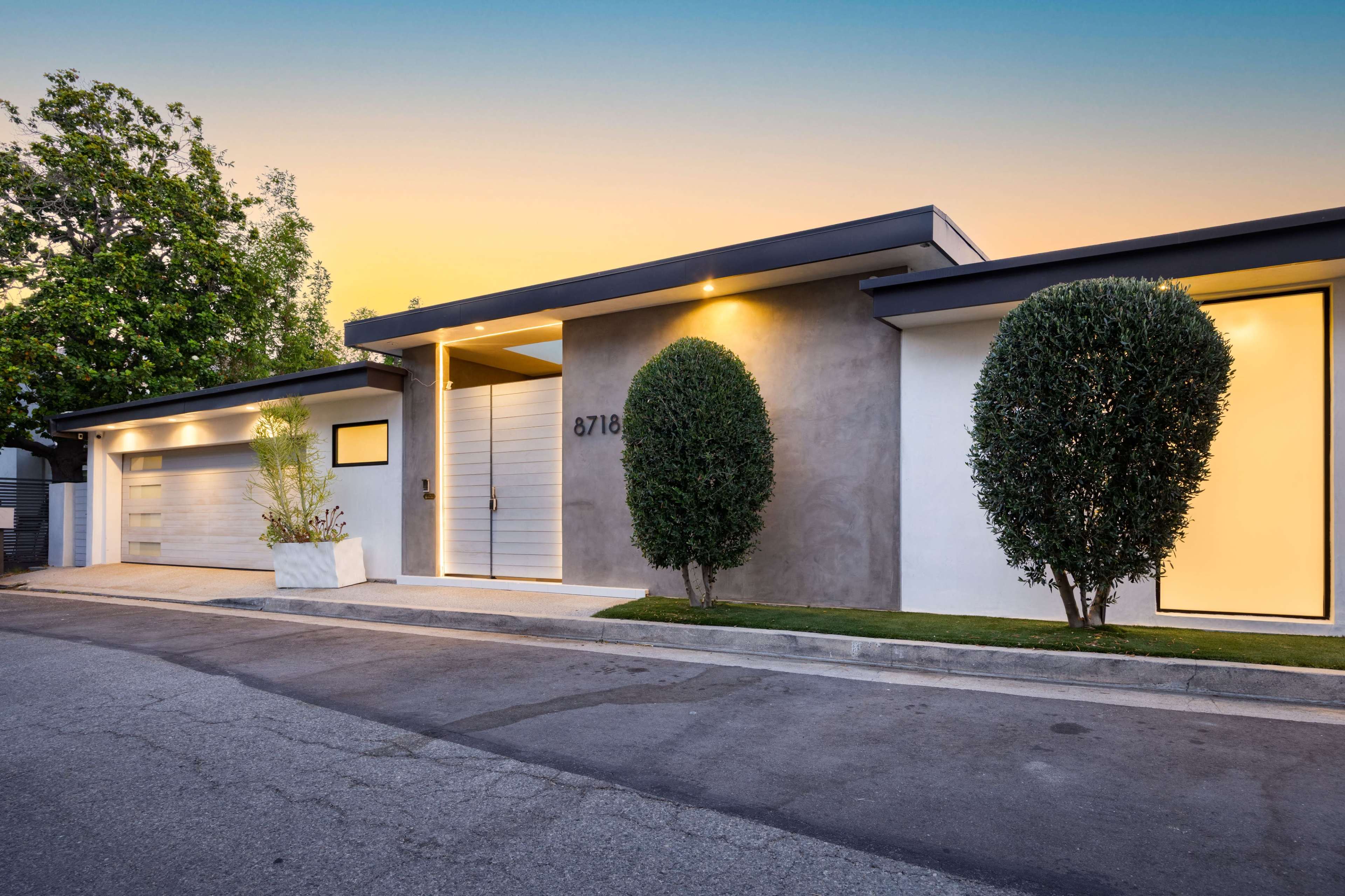 The image shows a modern single-story house with a sleek design, featuring two rounded bushes flanking the entrance and a driveway lined with a concrete path.