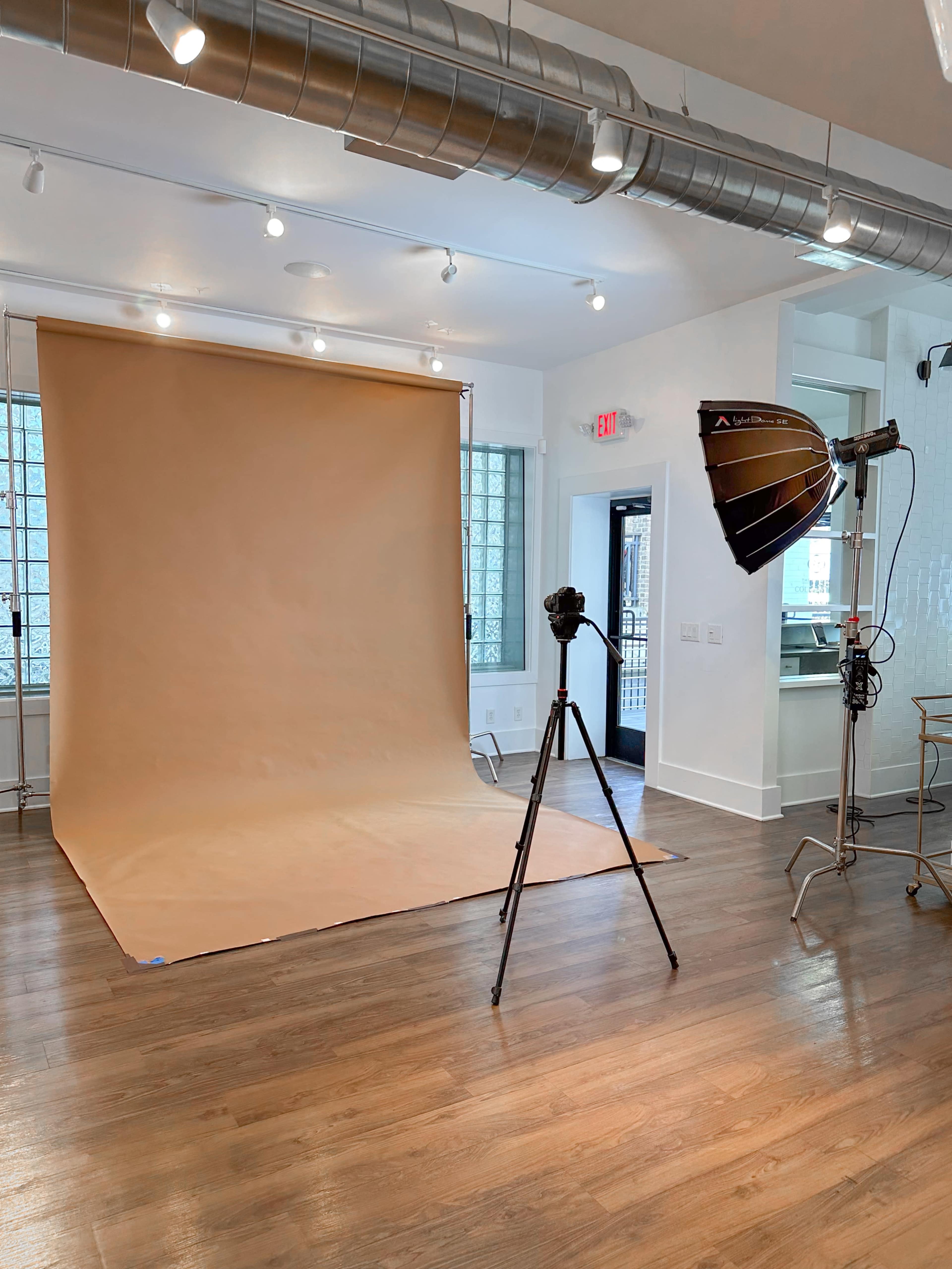 The image shows a photography studio with a beige backdrop, a camera tripod, and a lighting setup.